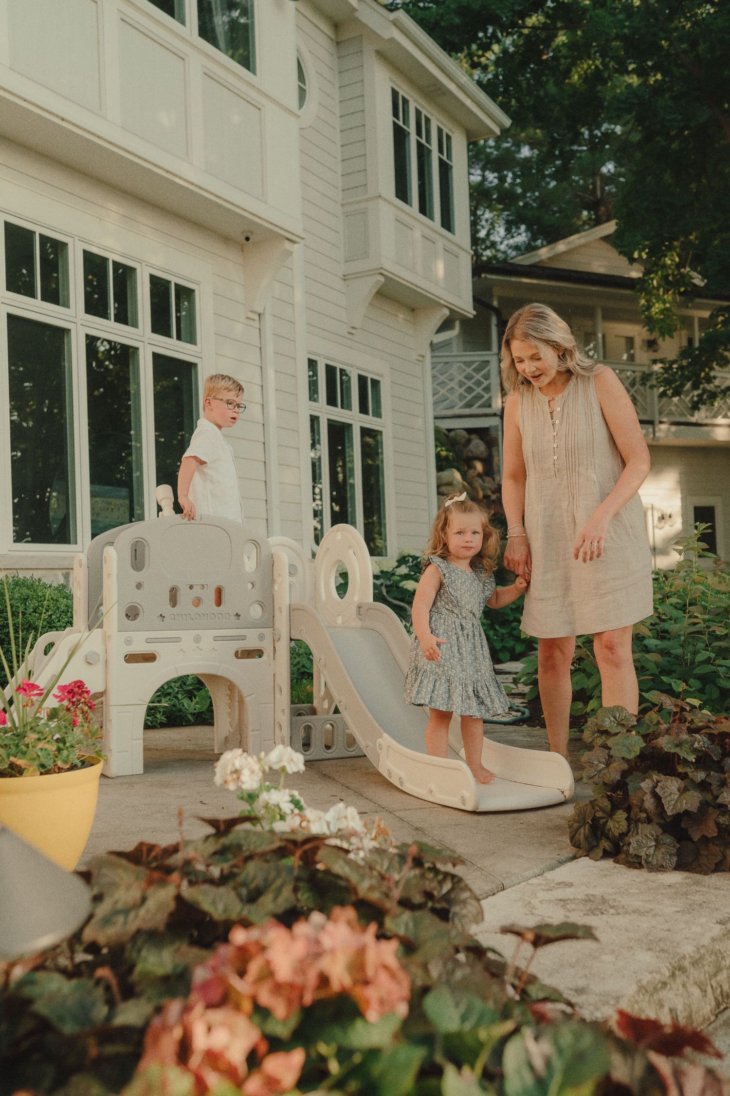 Smiling mother watches children play on backyard slide outside large white home.