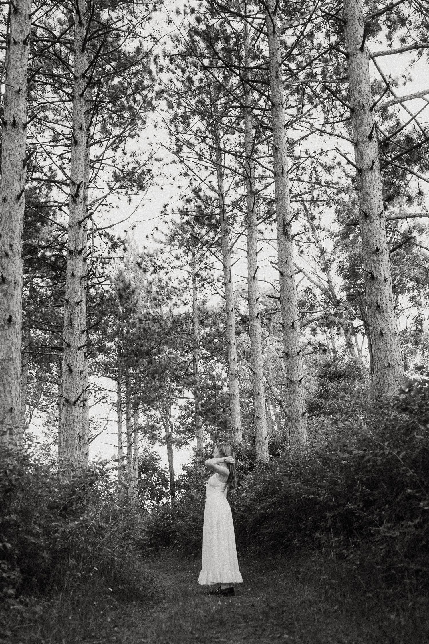 Woman in white dress standing among tall pine trees on forest path, black and white fine art photo.