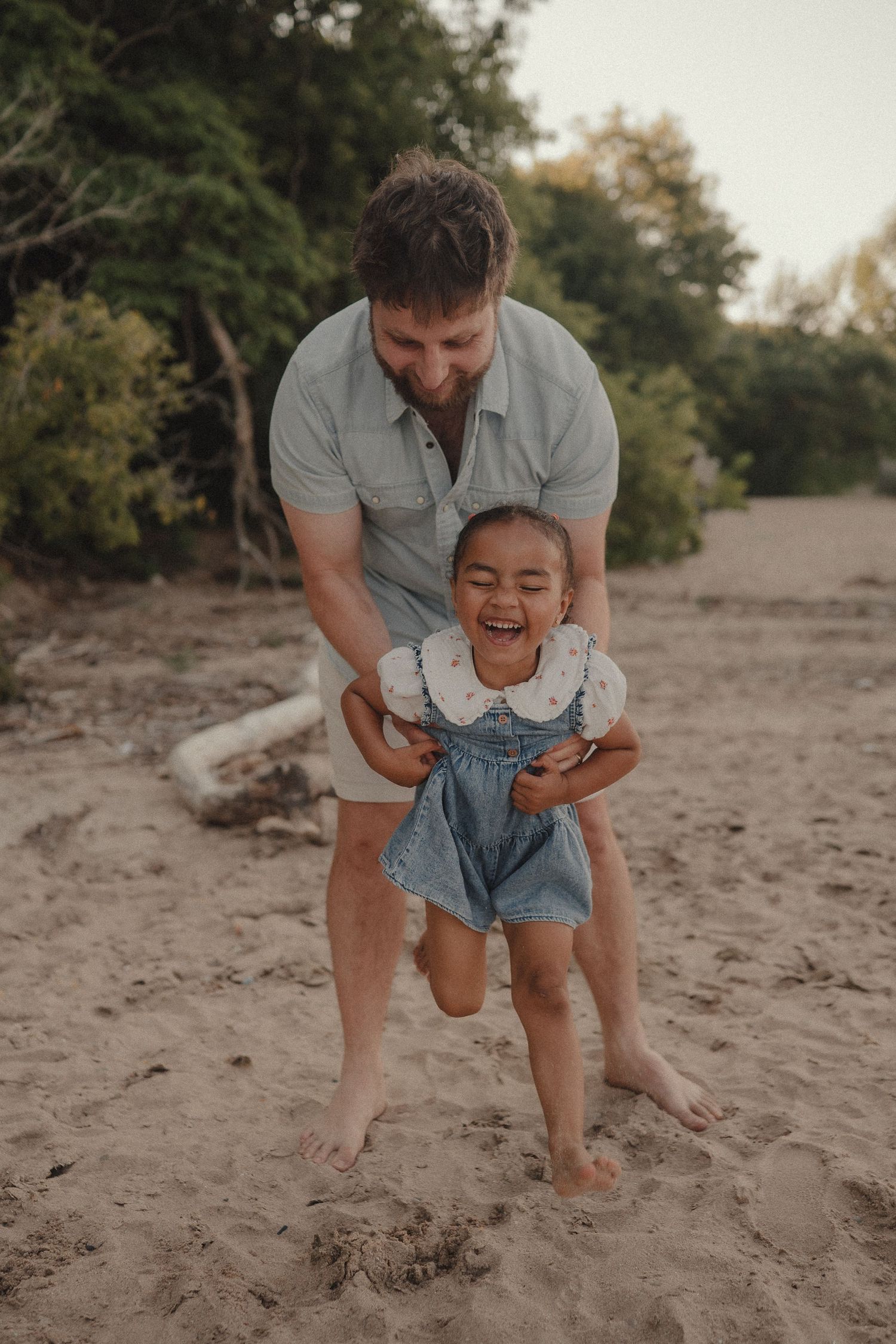 A laughing toddler girl in denim overalls plays on a sandy beach with her bearded father.