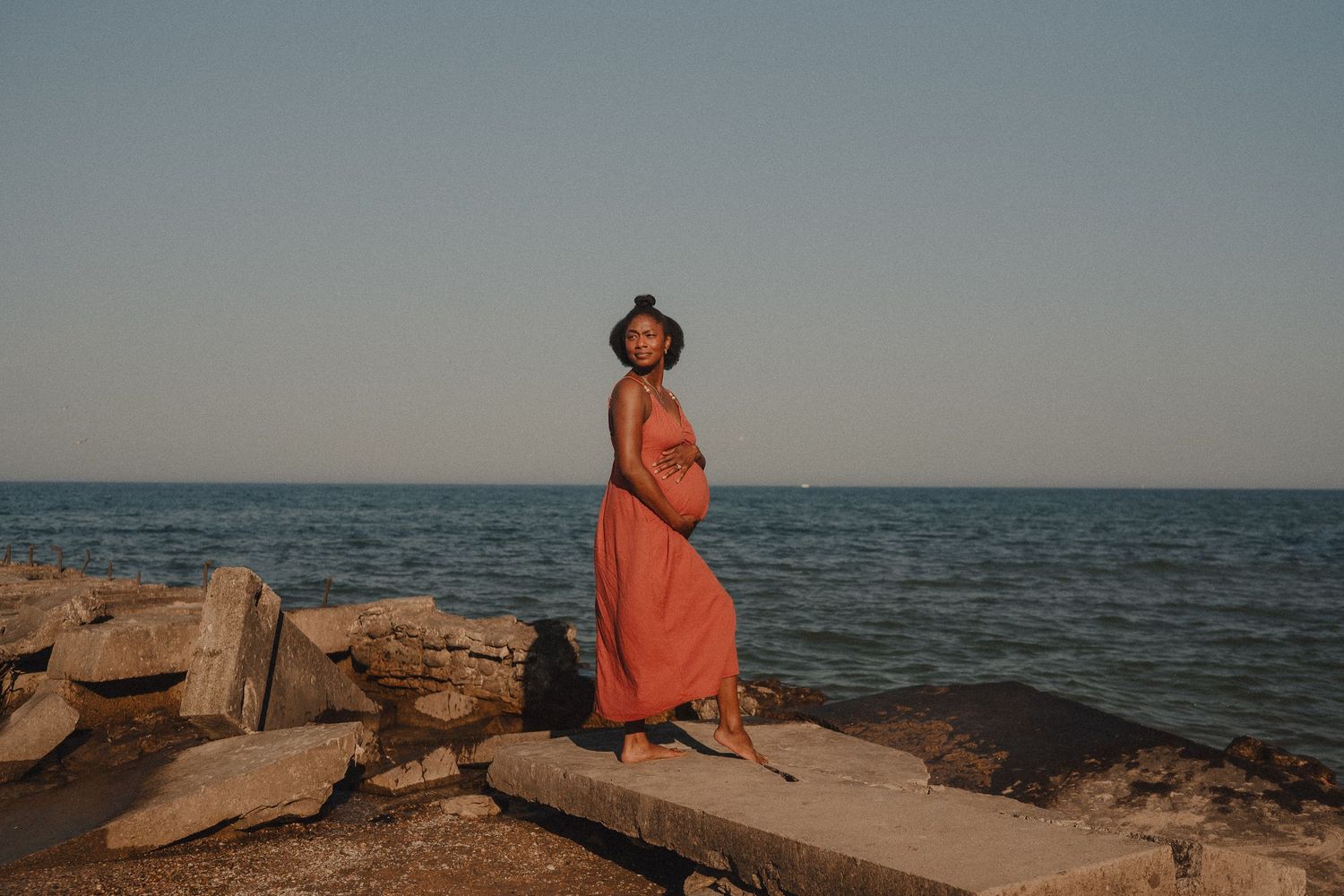 Pregnant woman in coral dress and hat standing on rocky shoreline at golden hour with ocean backdrop.