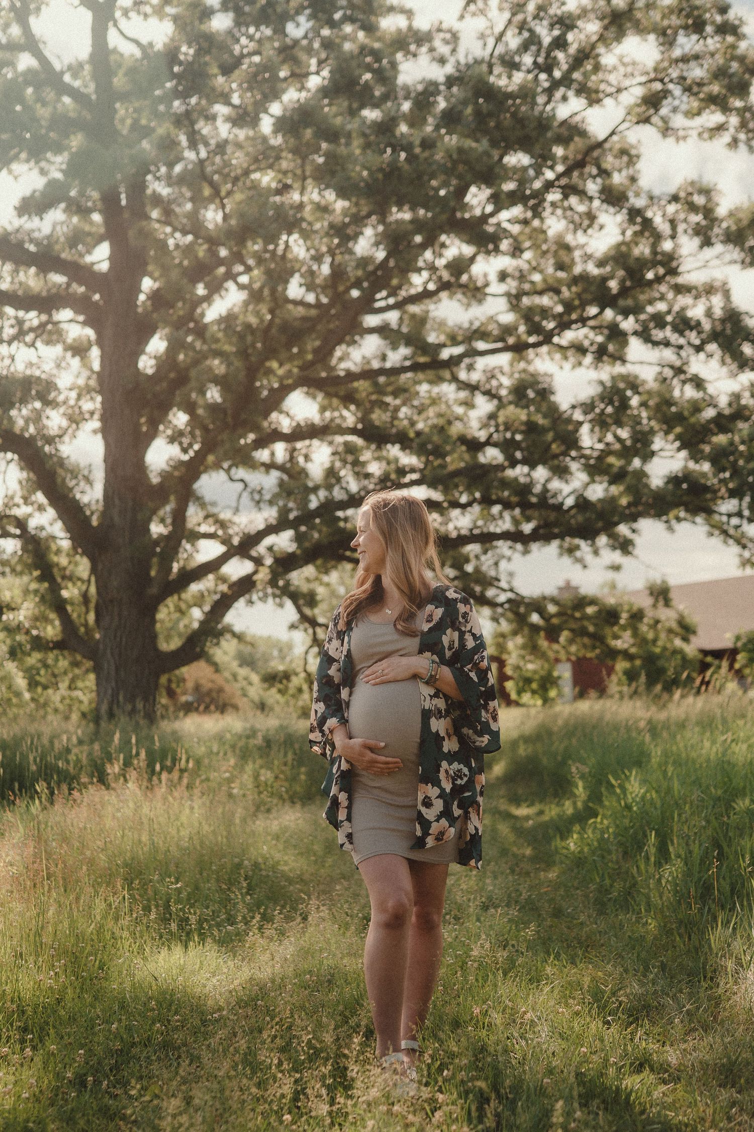 Pregnant woman in floral kimono cradling her baby bump in a golden-lit meadow with oak tree.