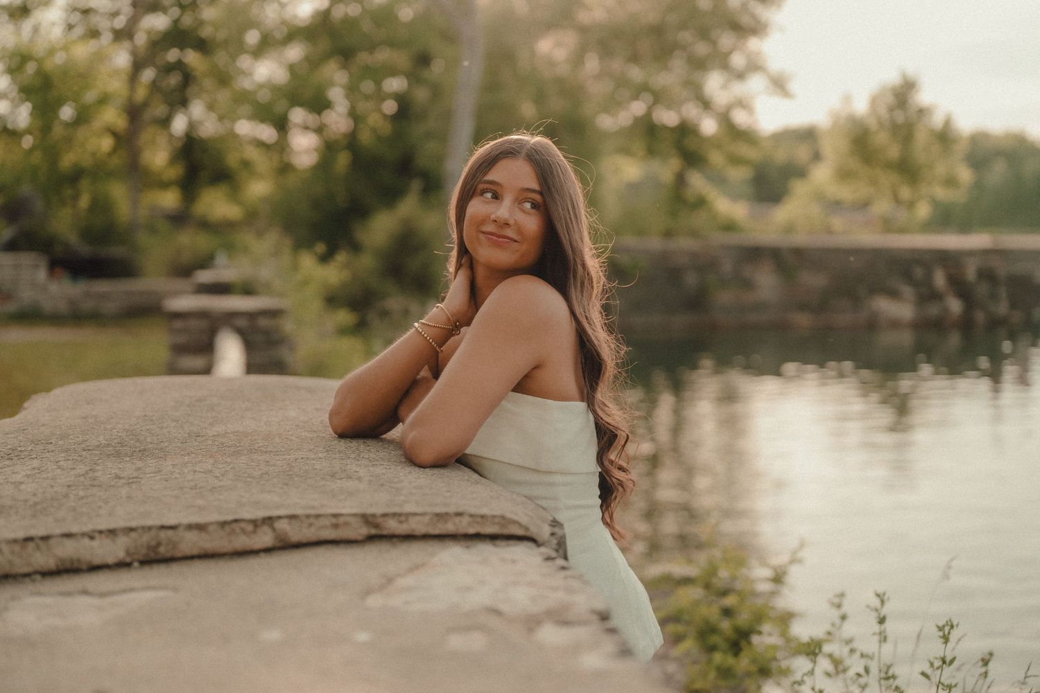 Young woman in white dress smiling by a serene lakeside, surrounded by lush greenery at golden hour.