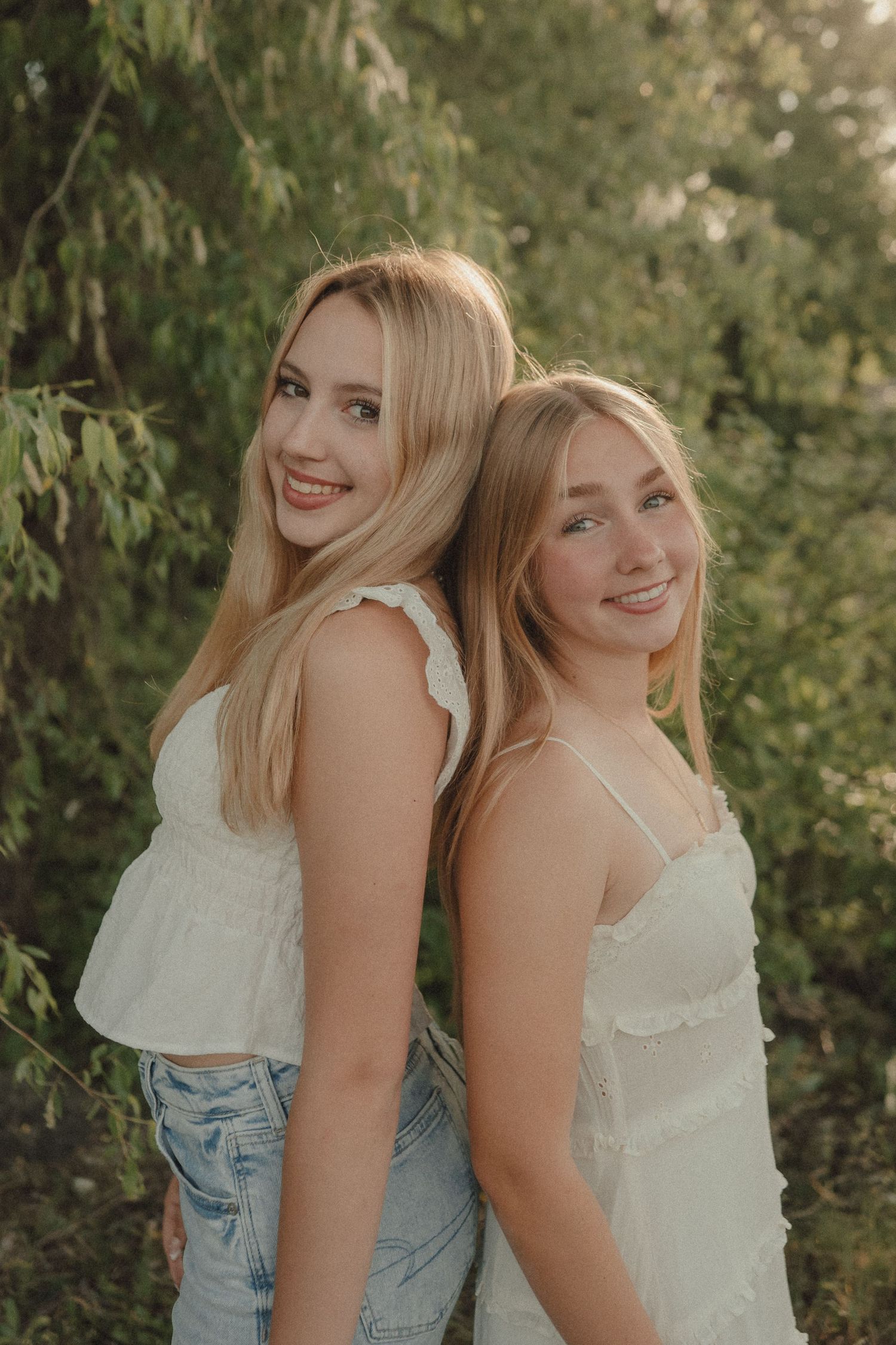 Two smiling blonde young women in white tops pose back-to-back outdoors among green trees in warm sunlight.
