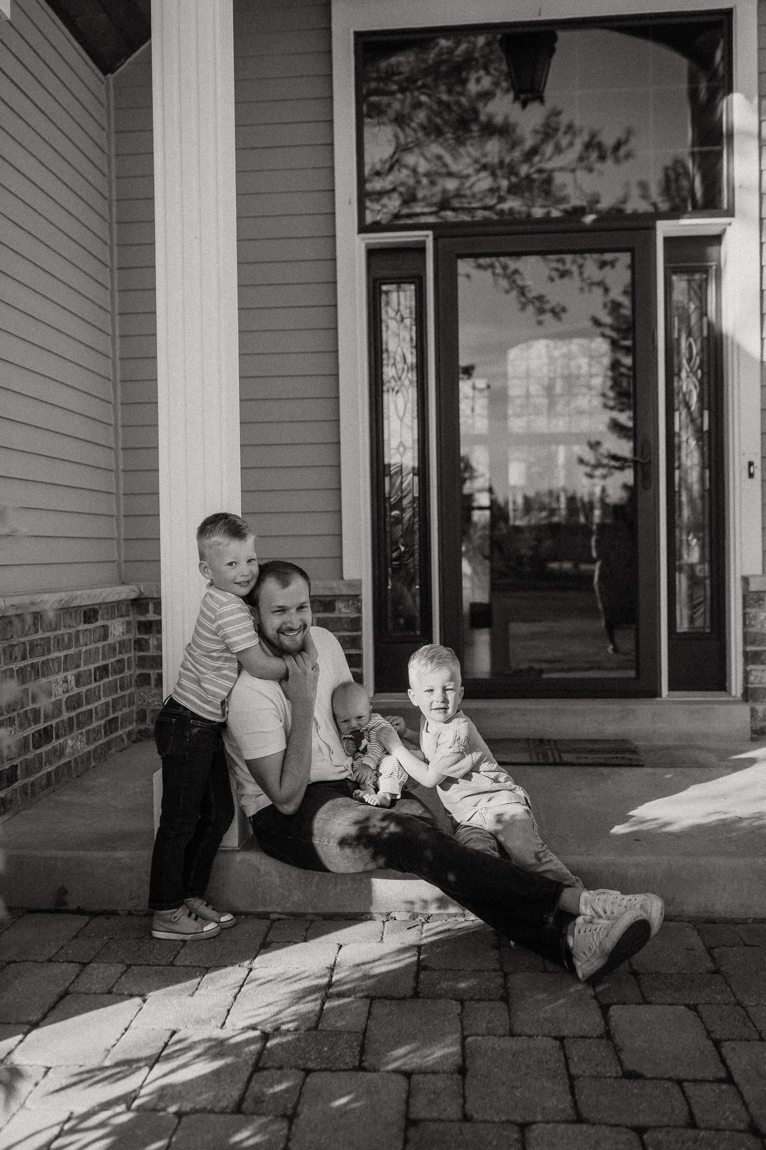 Black and white photo of a father sitting on front porch steps with two young sons smiling.