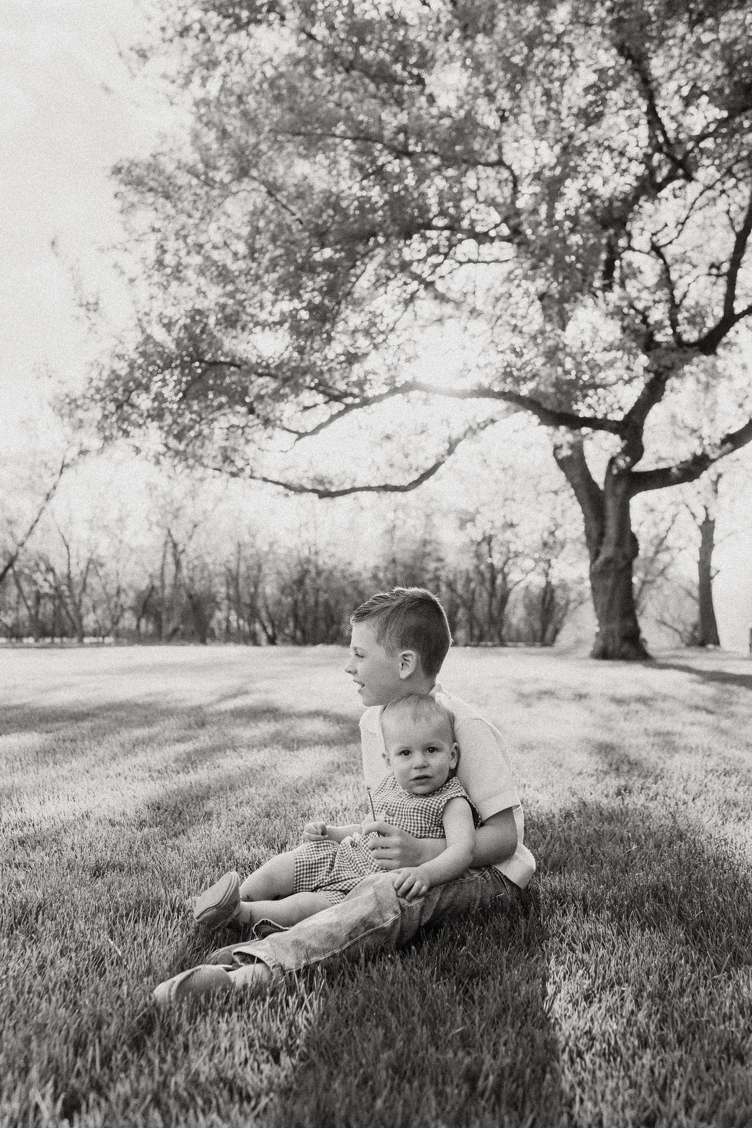 Black and white photo of two young siblings sitting together on grass beneath a large tree.