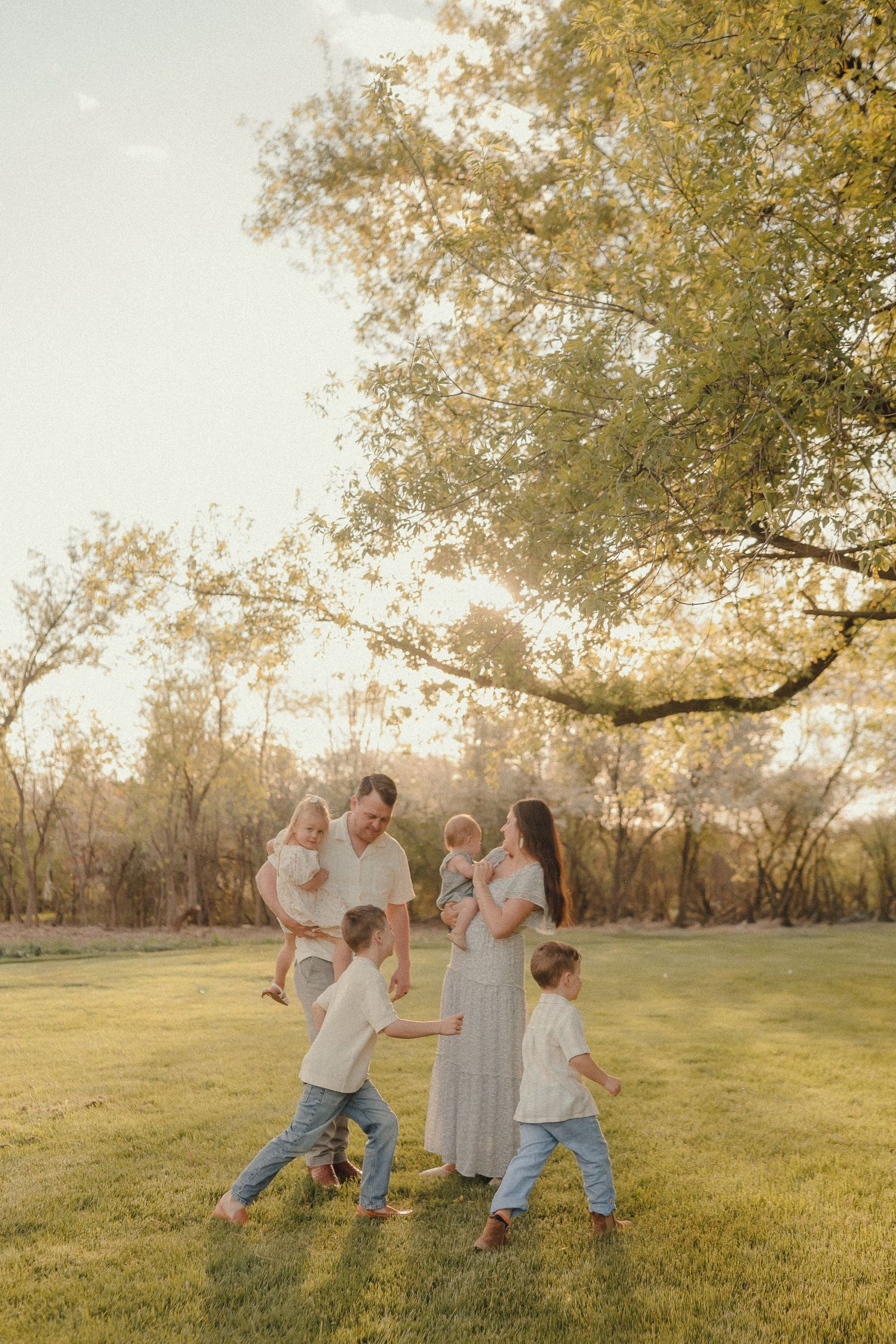 Family of five playing together on a green lawn at golden hour beneath a large sprawling tree.
