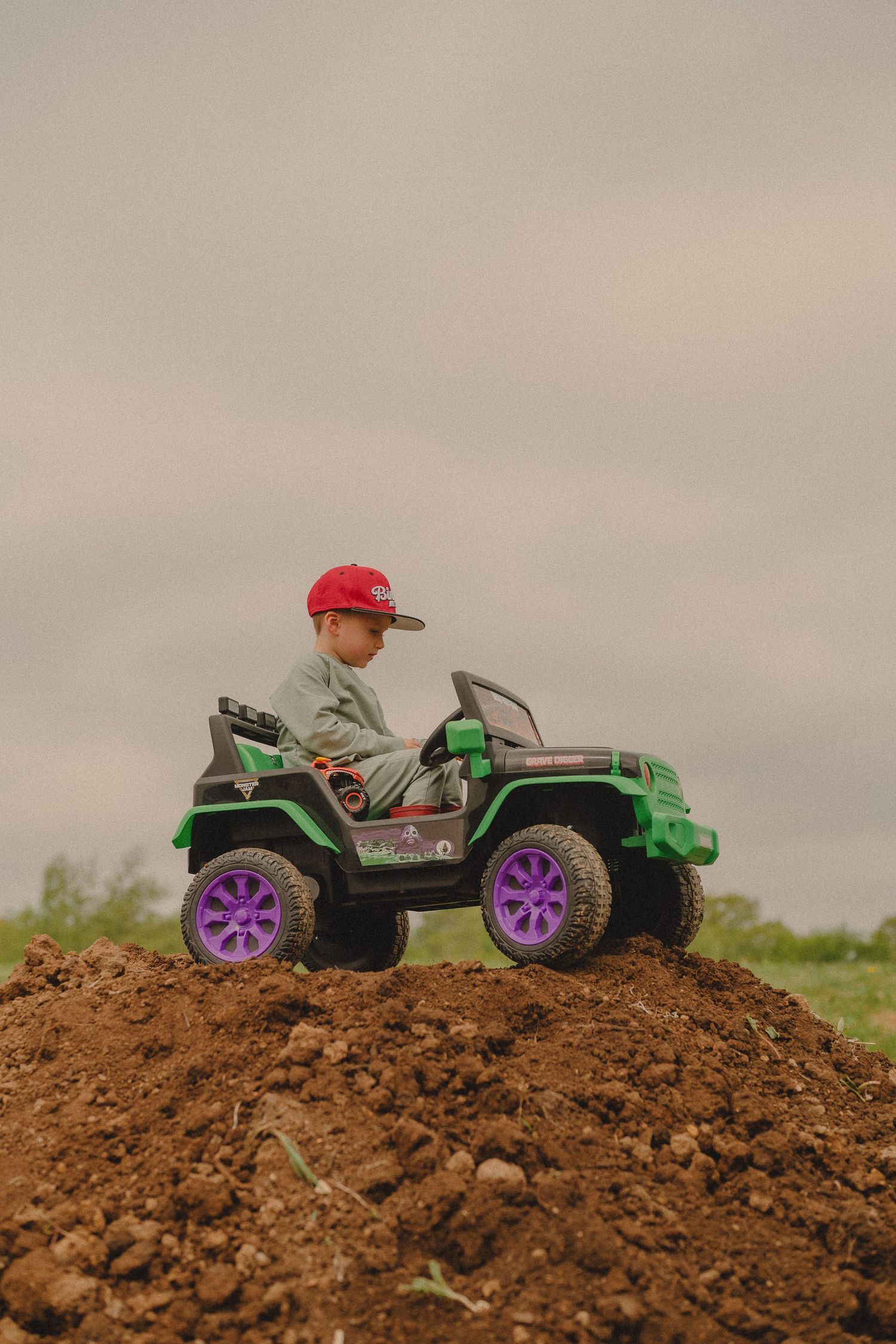 Young child in red snapback cap rides green monster truck toy on outdoor dirt pile.