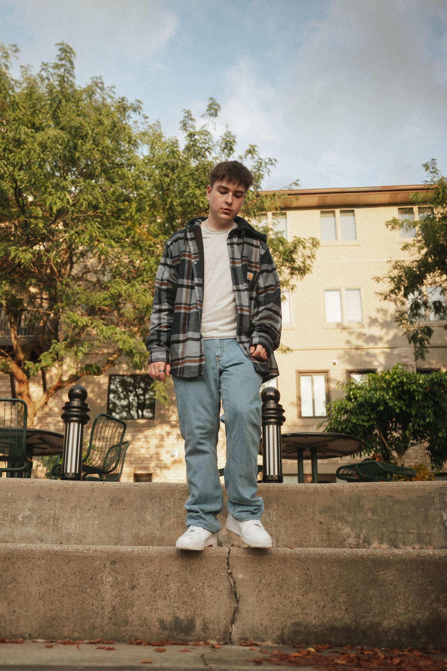 A person in a plaid shirt and jeans sits casually on a concrete wall on an autumn day with fallen leaves below.