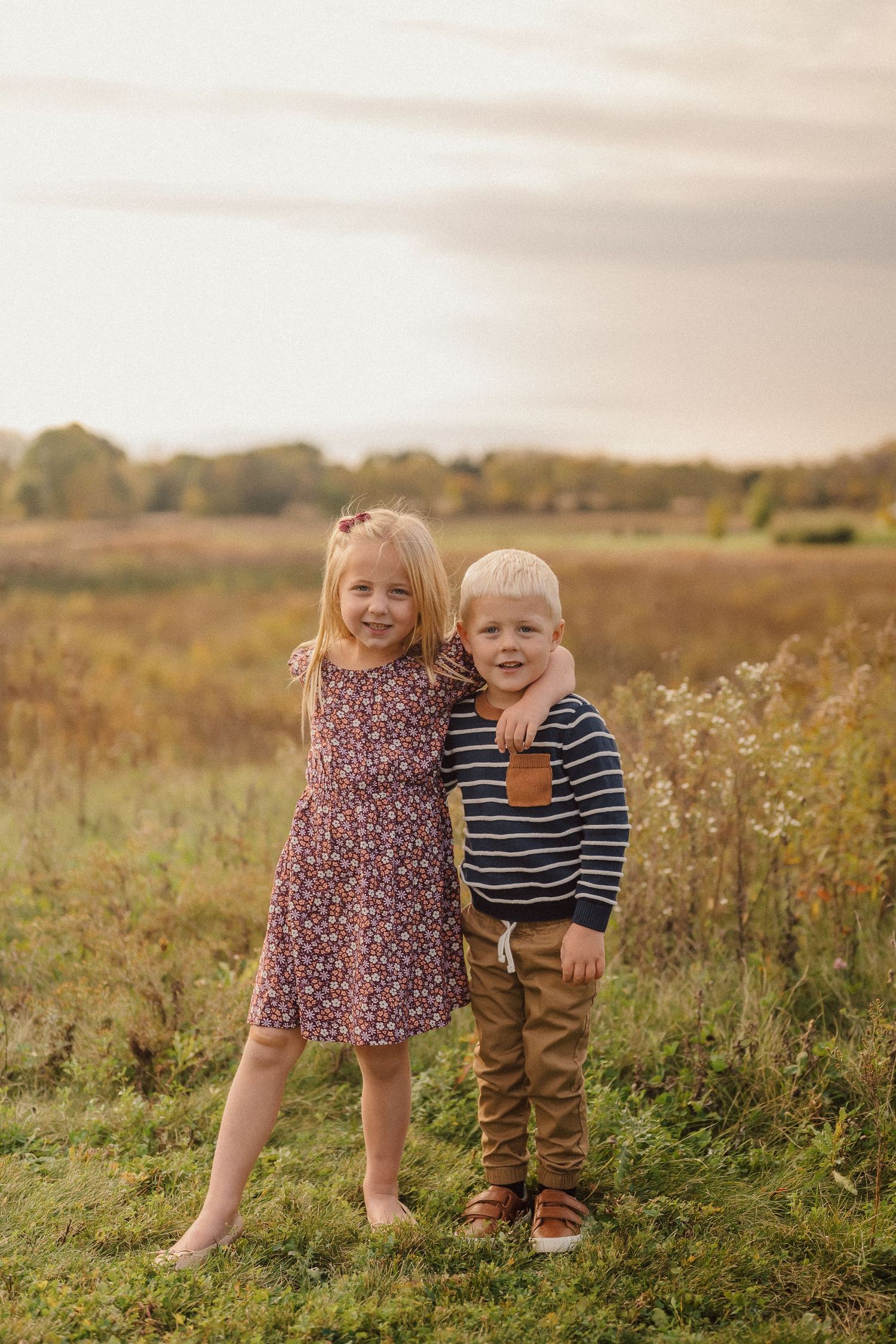 Two small children stand together in a grassy meadow during golden hour.