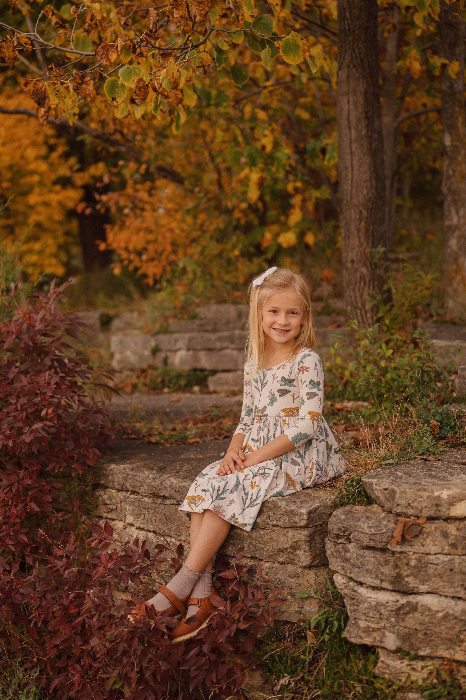 A child in a floral dress sits on a rustic stone wall surrounded by vibrant fall foliage.