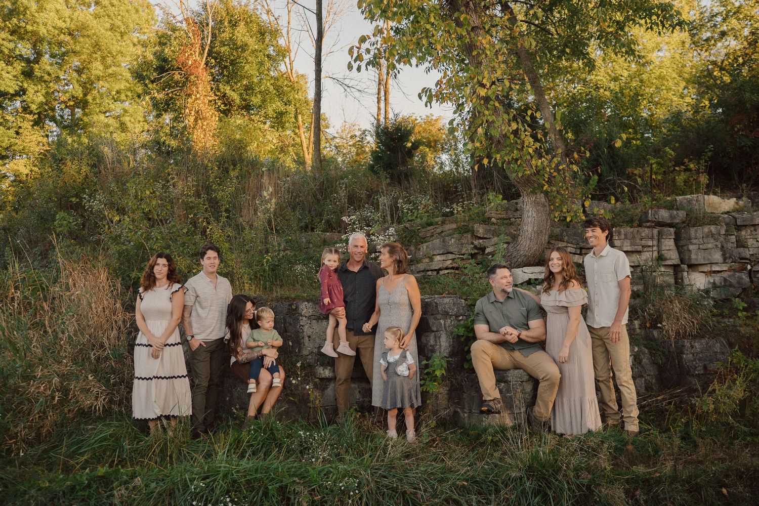 A large family group poses together outdoors at sunset against a rustic stone wall background.