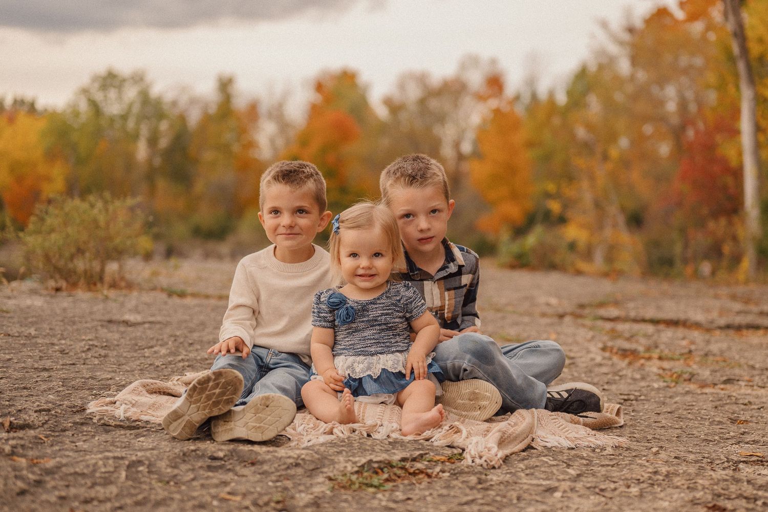 Children sit together on rocky ground surrounded by warm autumn foliage in the background.