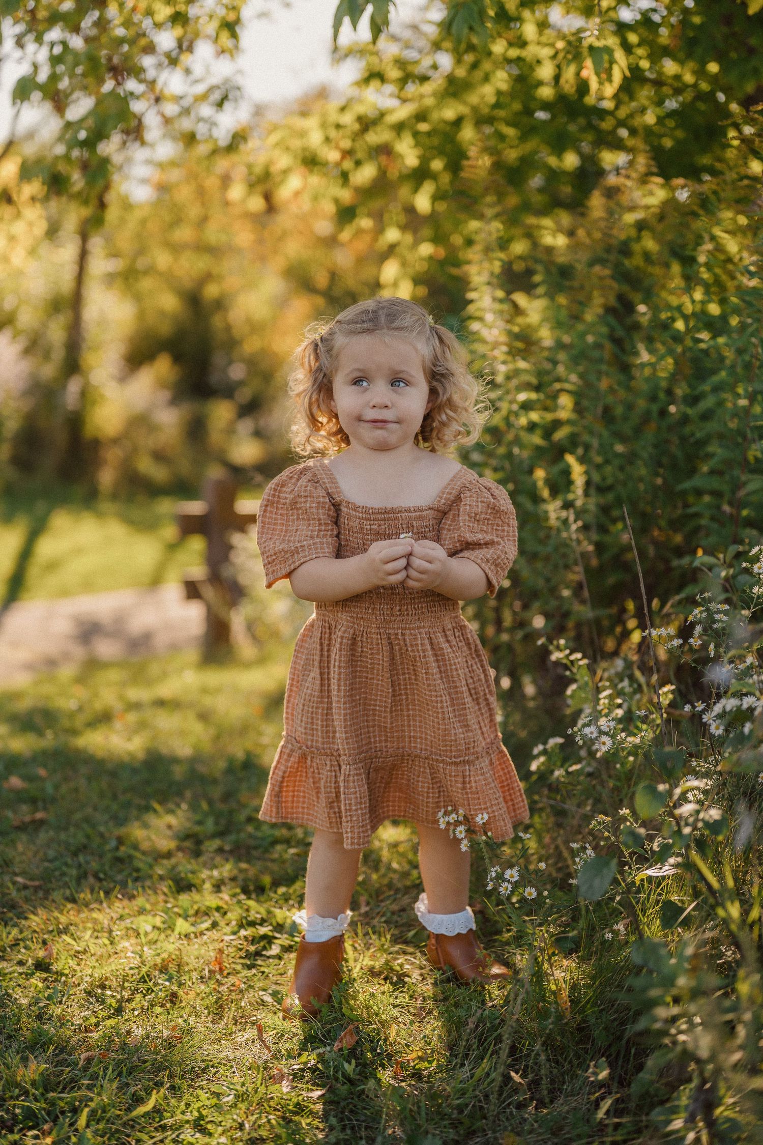 Child in brown dress poses outdoors among green foliage in natural sunlight.