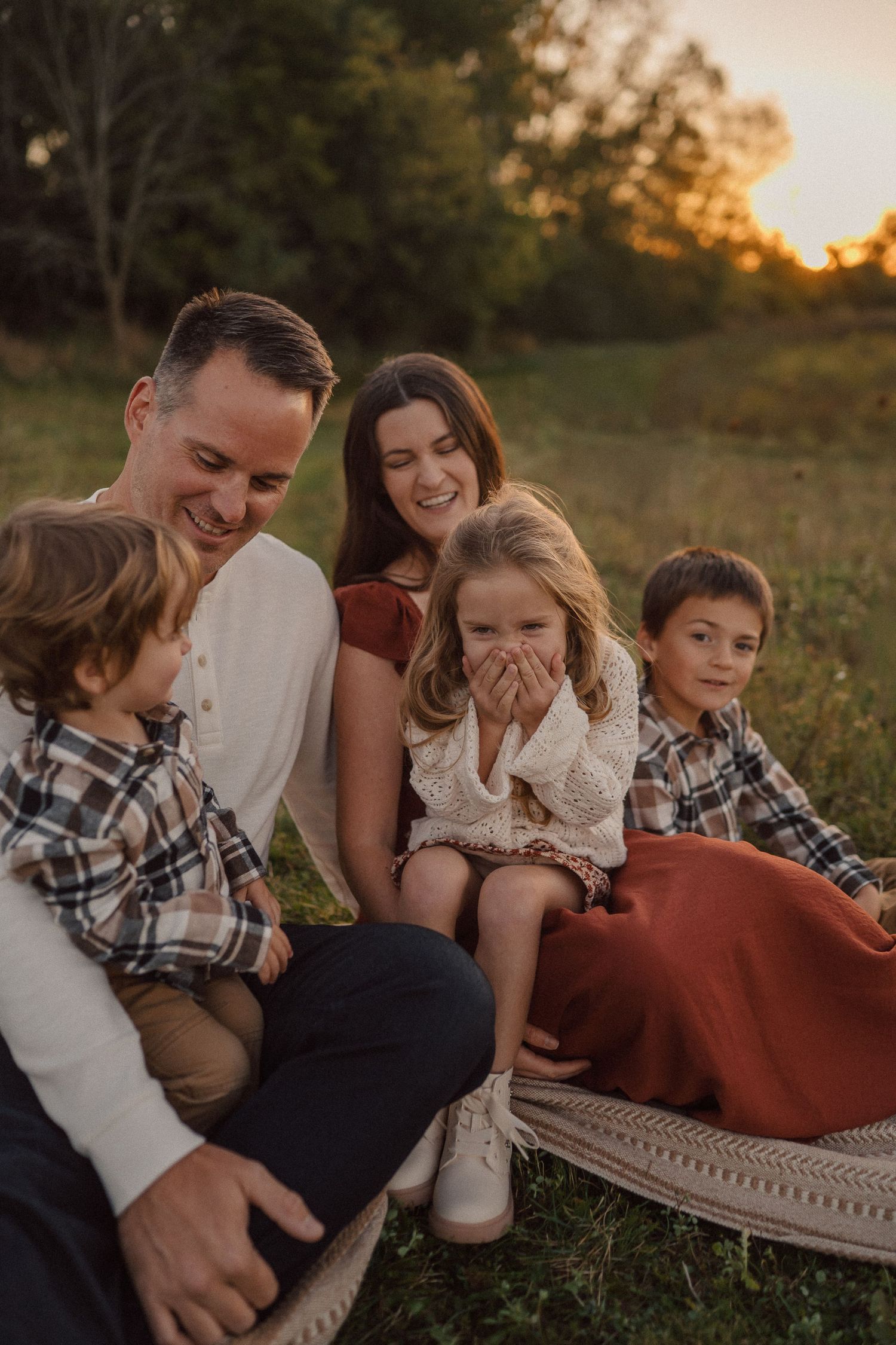 Family relaxes together on blanket in grassy field during golden sunset photo session.