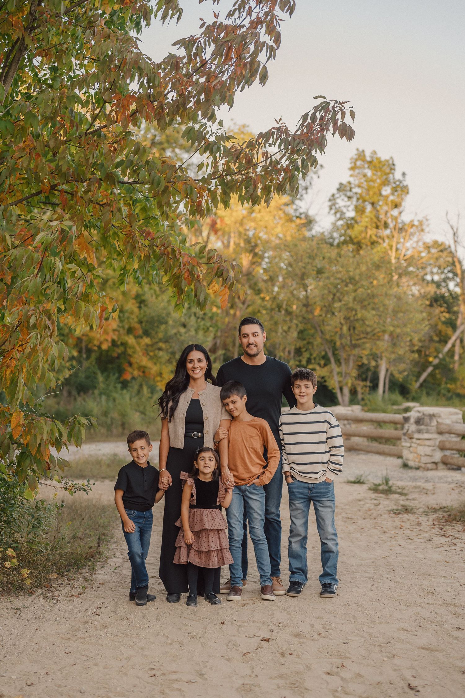 Family group stands together on sandy ground with autumn trees and foliage in the background.