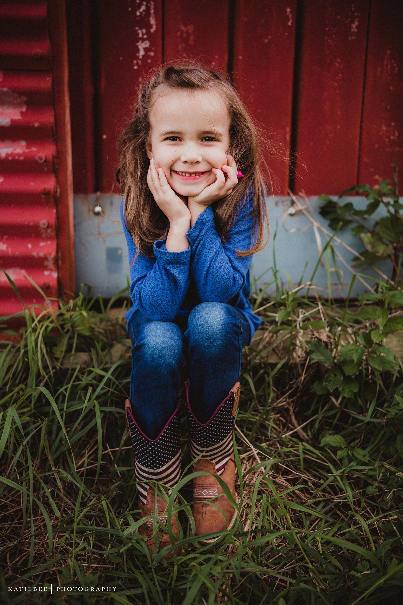 Portrait of a child in a blue sweater sitting in grass against a red barn wall.