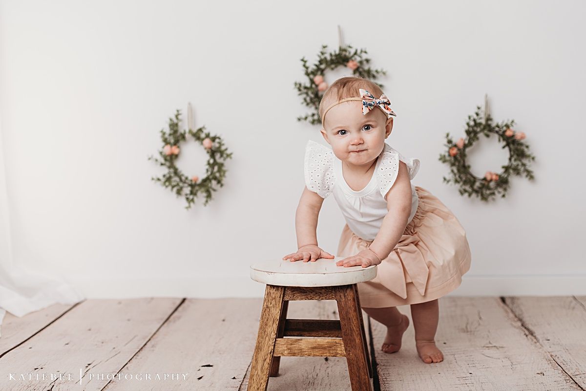 Baby in floral crown sitting near wooden stool against white wall with decorative wreaths.