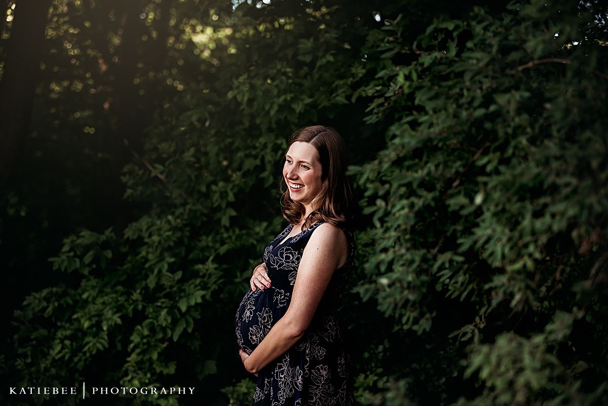 An expectant mother in a patterned dress stands in dappled sunlight against leafy foliage.