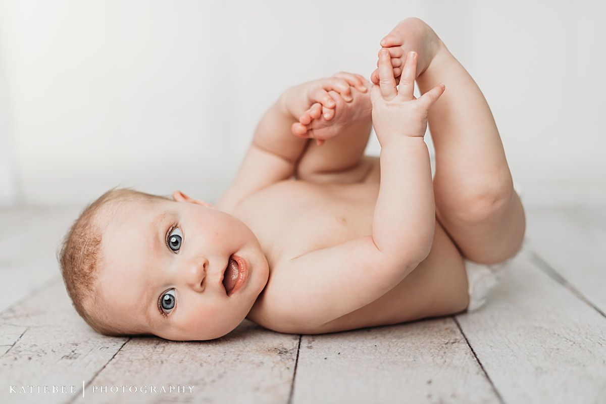 Baby lying on white wooden floor grabbing feet during portrait session.