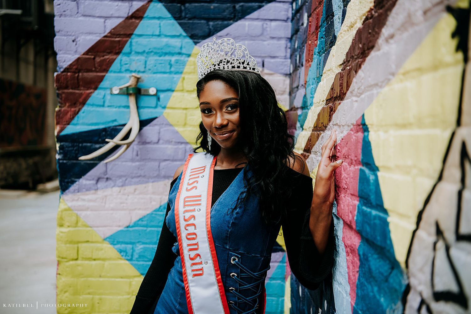 Portrait in front of vibrant geometric street art mural wearing a sash and crown.