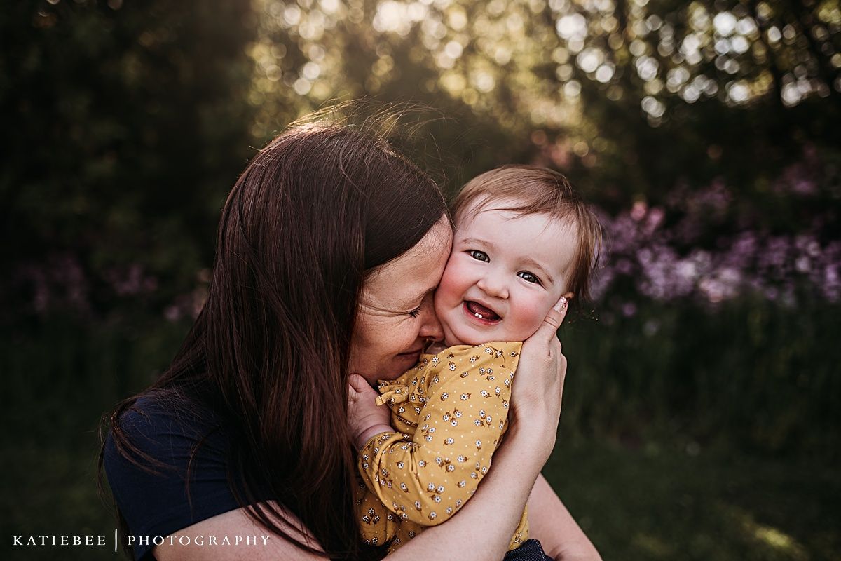 Tender moment captured outdoors with sunlit trees and purple wildflowers in the background.