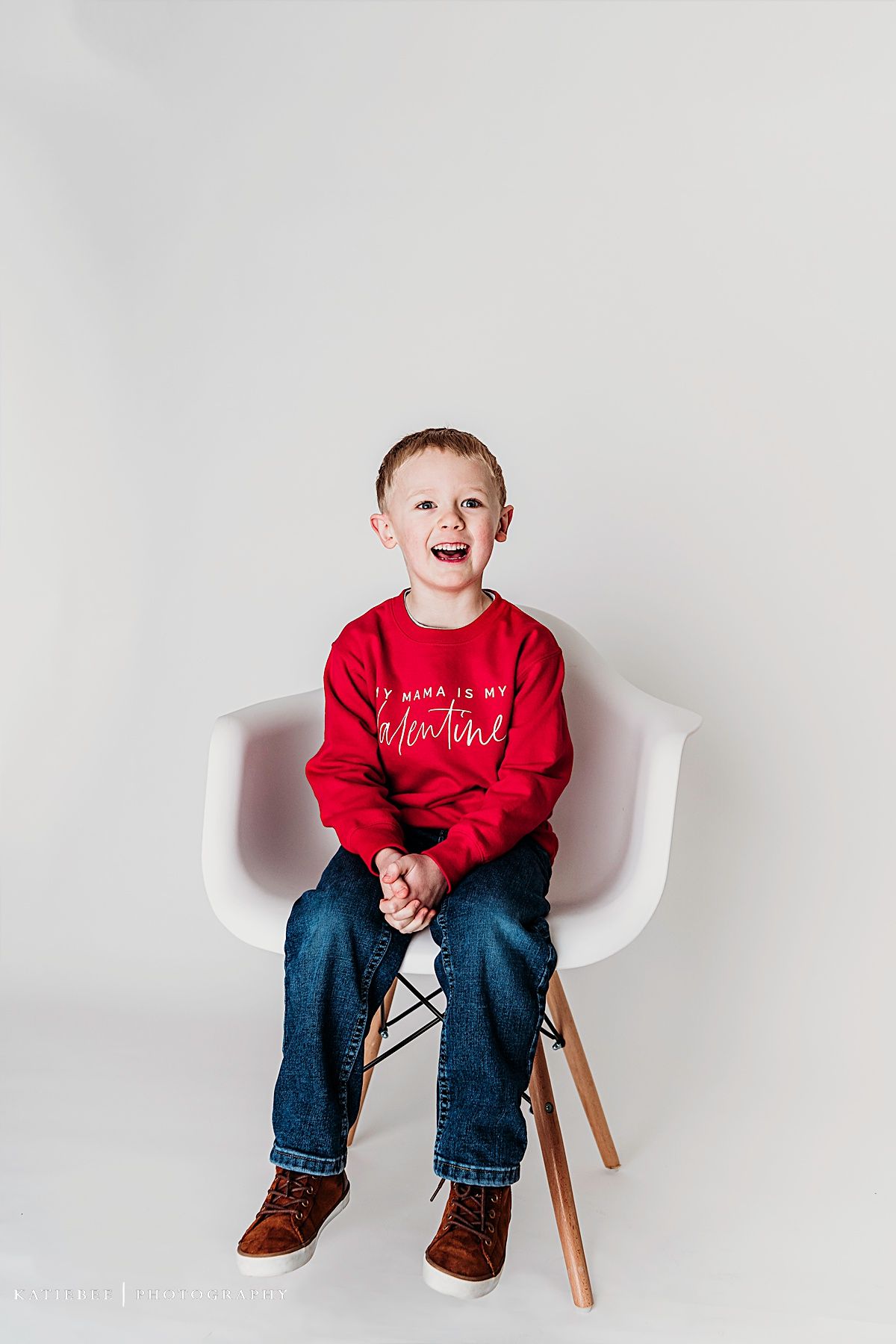Joyful pose on white chair wearing red sweater with valentine message and blue jeans.