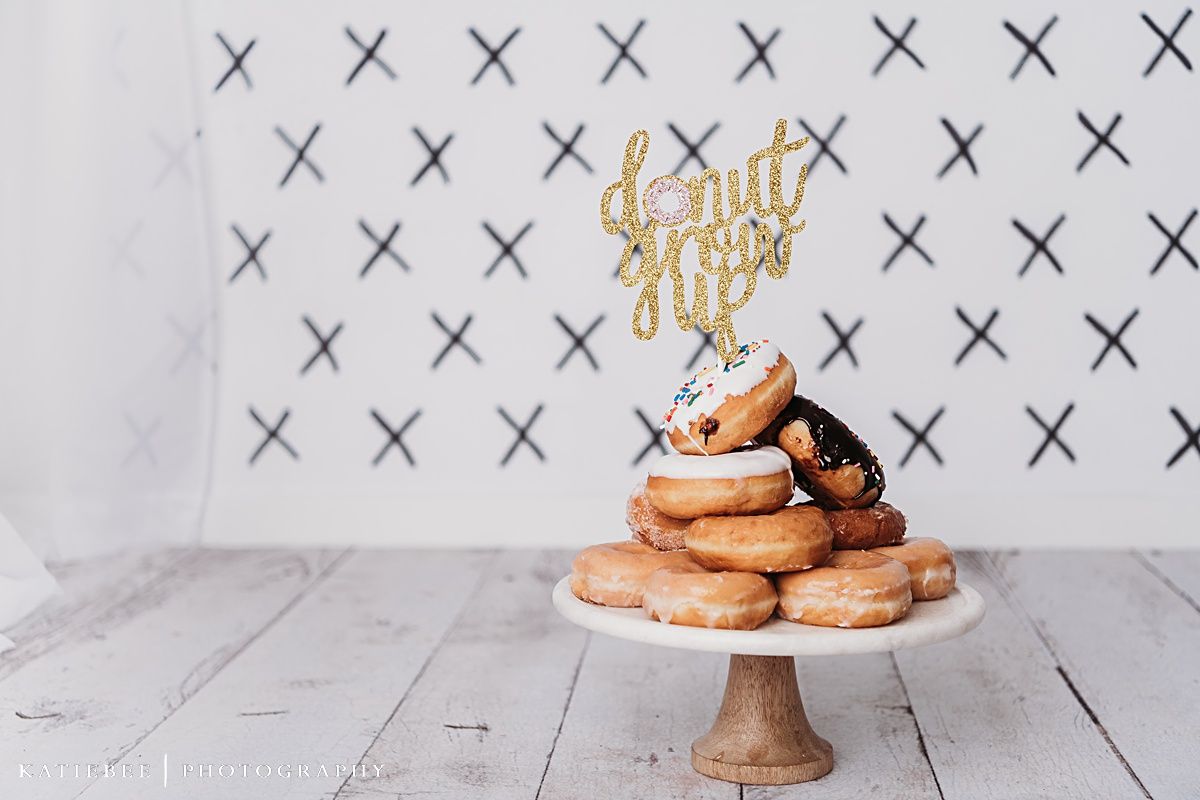 Cake stand displays stacked donuts with gold cake topper against white X-patterned backdrop.