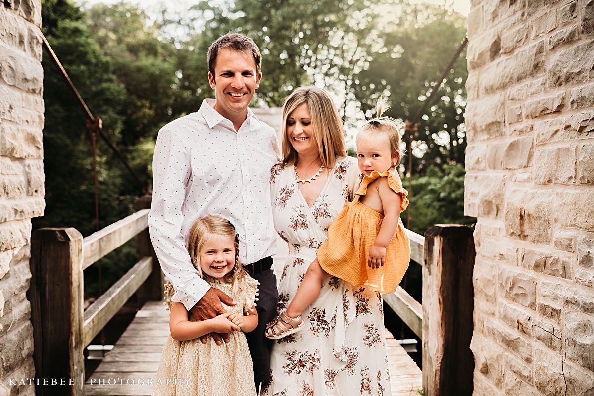 Family stands together on a wooden bridge wearing coordinated white and floral outfits.