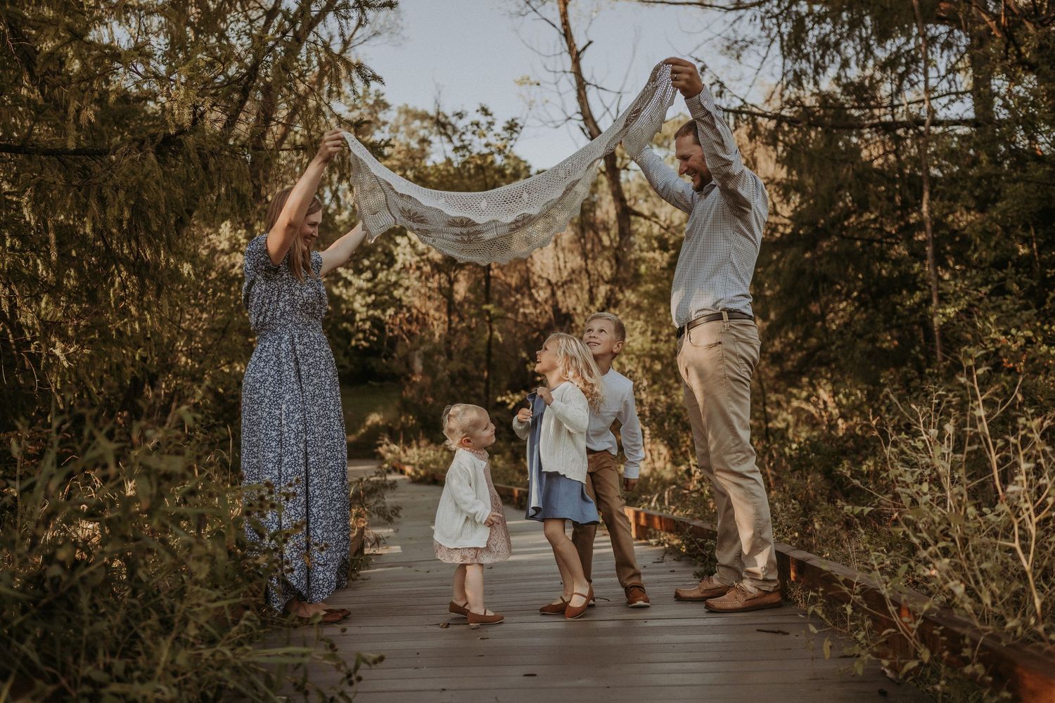 A family plays together on a wooden boardwalk, holding up a white sheet.