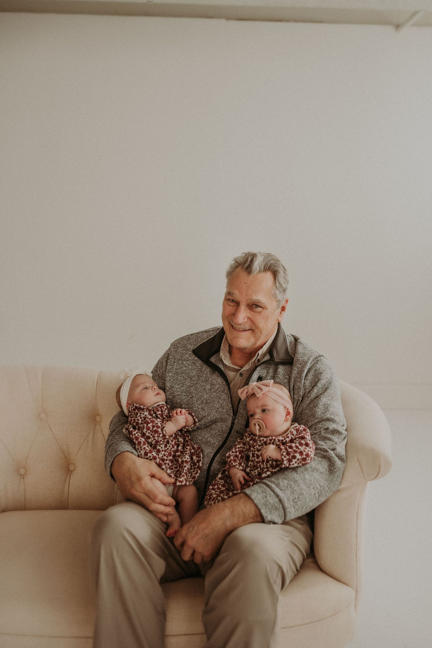 A person in a grey sweater holds two infants wearing matching floral outfits on a beige couch.