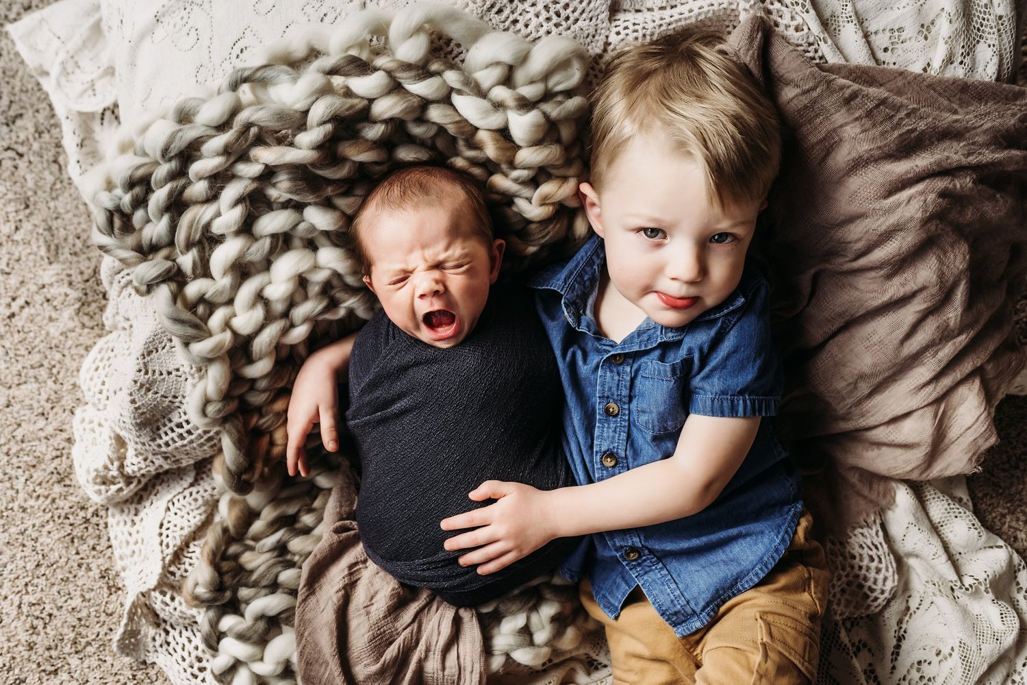Two siblings laying on a chunky knit blanket with contrasting expressions of joy and calm.