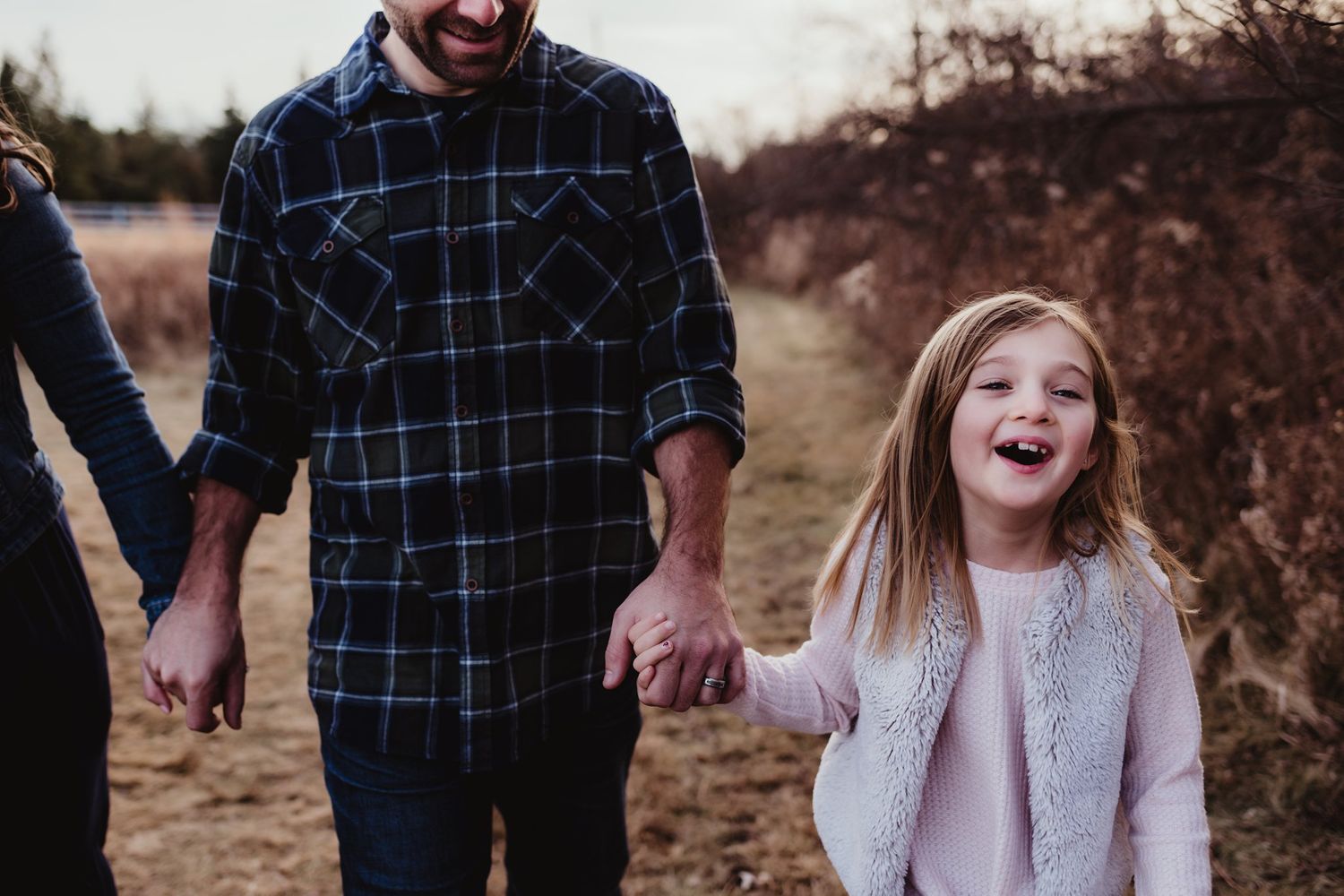 Family walking outdoors holding hands with little girl laughing in pink sweater.