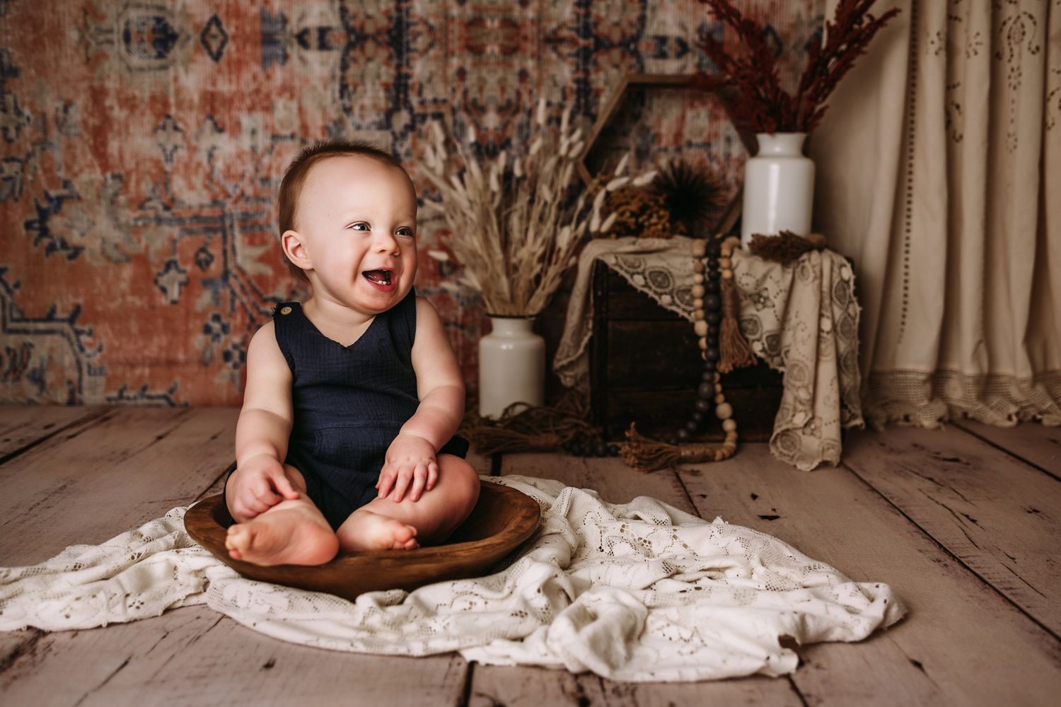 Happy baby in navy outfit sits in wooden bowl against rustic backdrop with dried flowers.