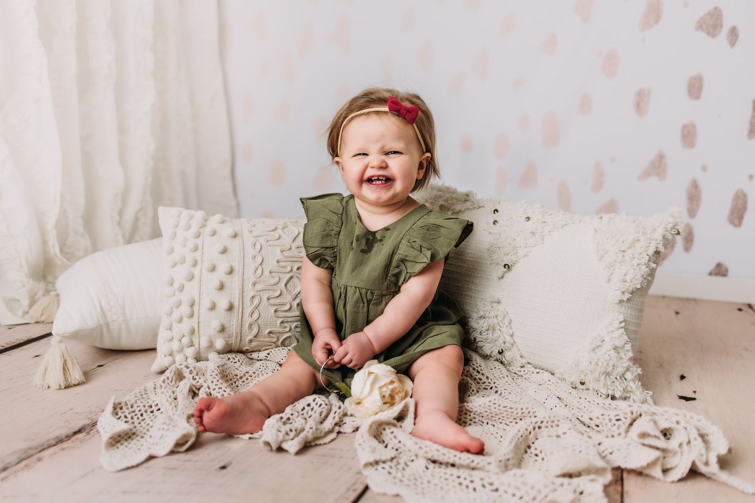 Happy baby in olive green outfit sits among cream knit blankets.