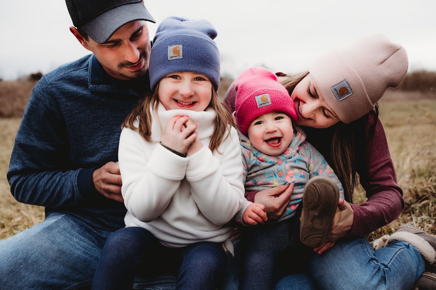 Candid outdoor family photo showing joyful expressions and winter hats on a cloudy fall day.