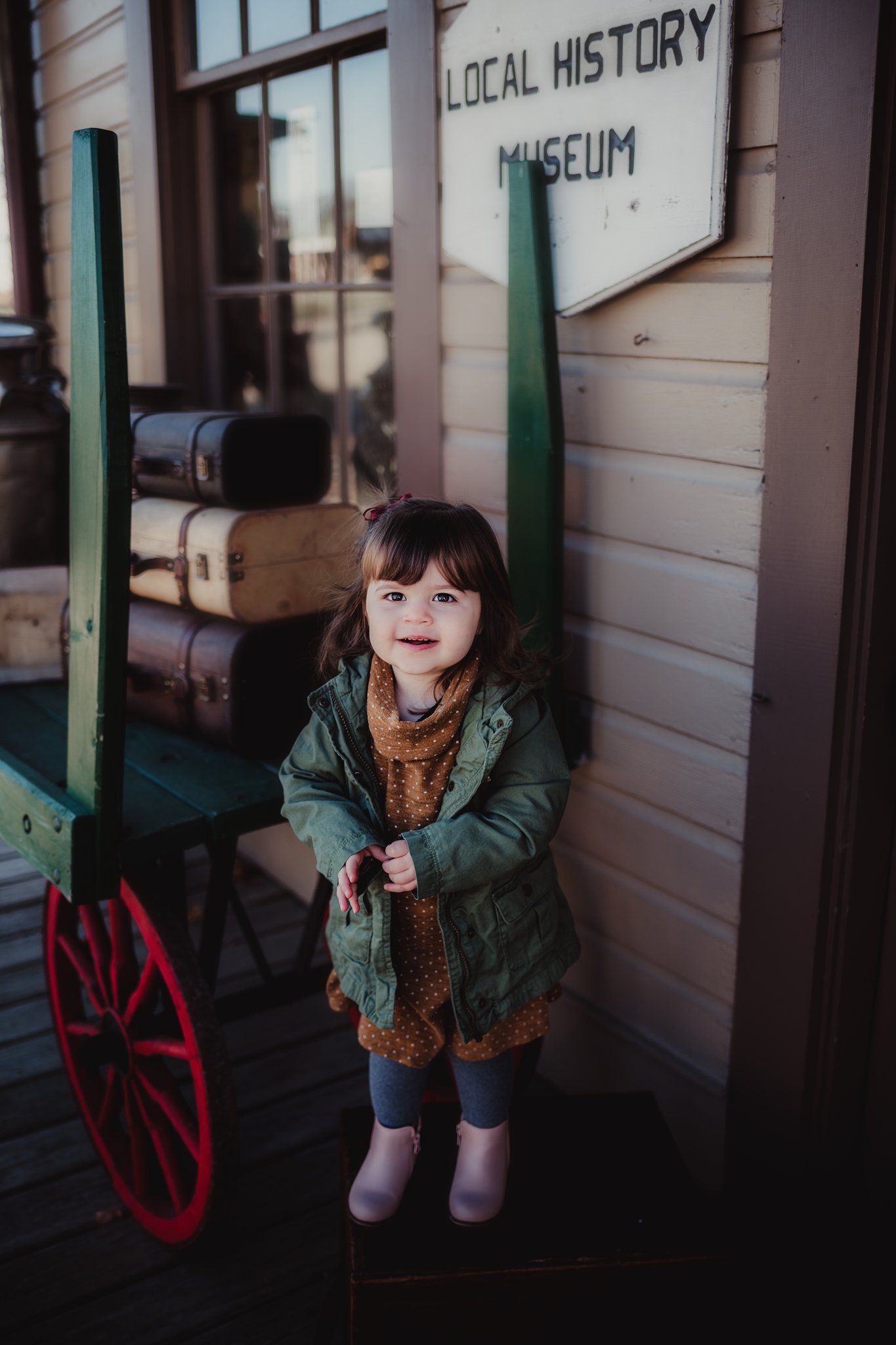 A small child stands outside a local history museum wearing a green jacket and polka dot dress.
