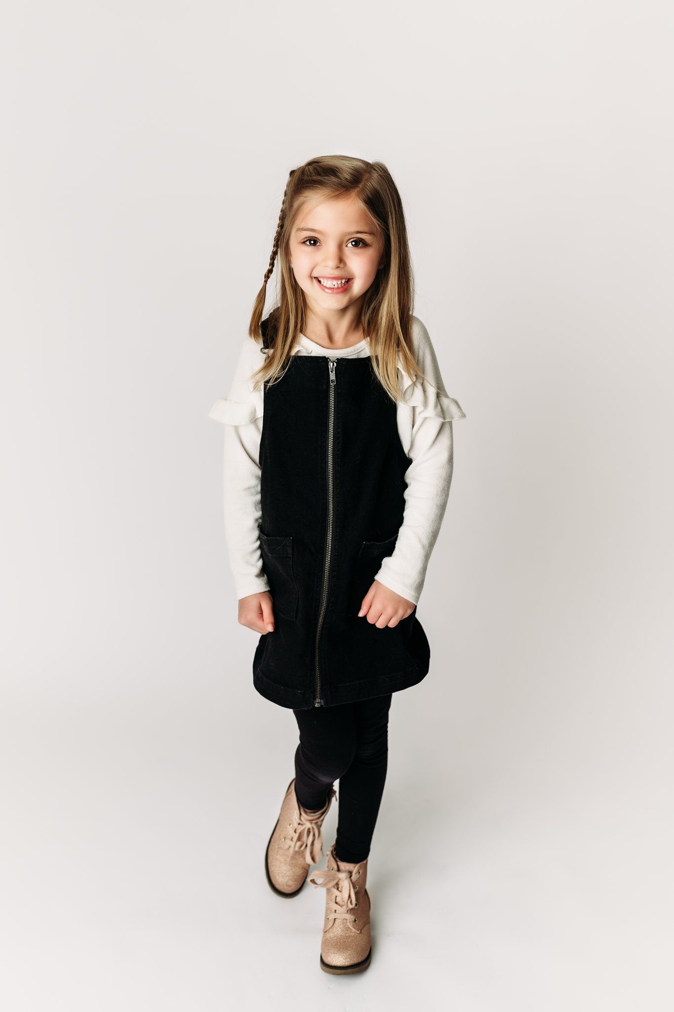 Young child wearing black jumper dress with white long sleeves and brown boots posing against white background.