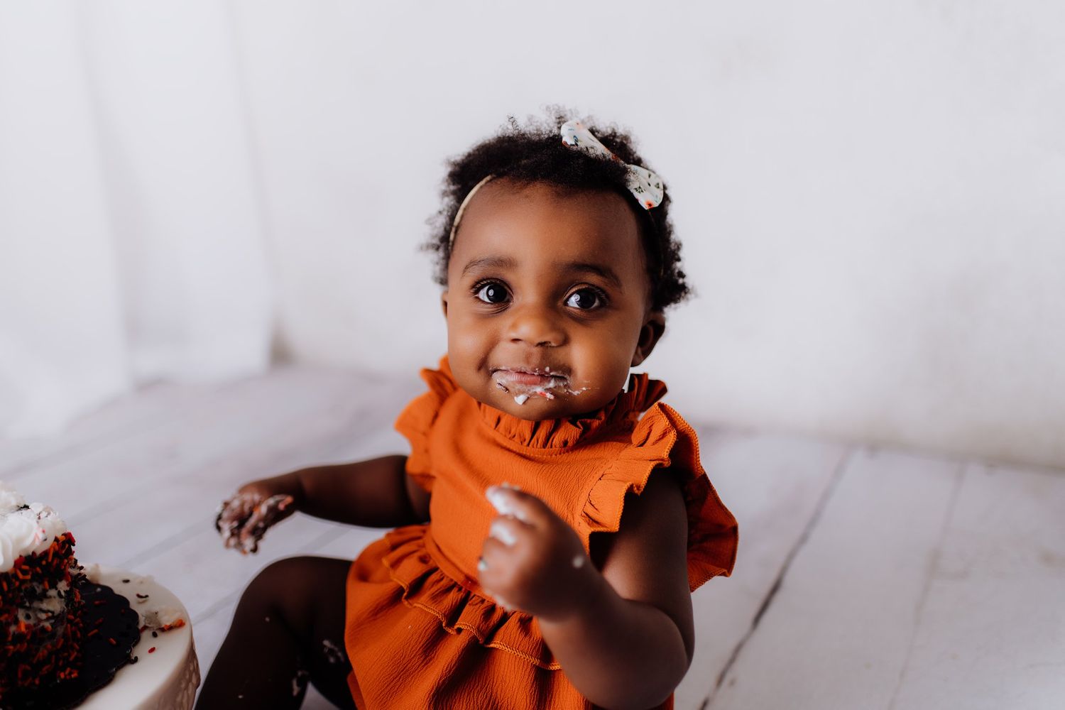 A toddler in an orange ruffled dress enjoys cake during a photoshoot.