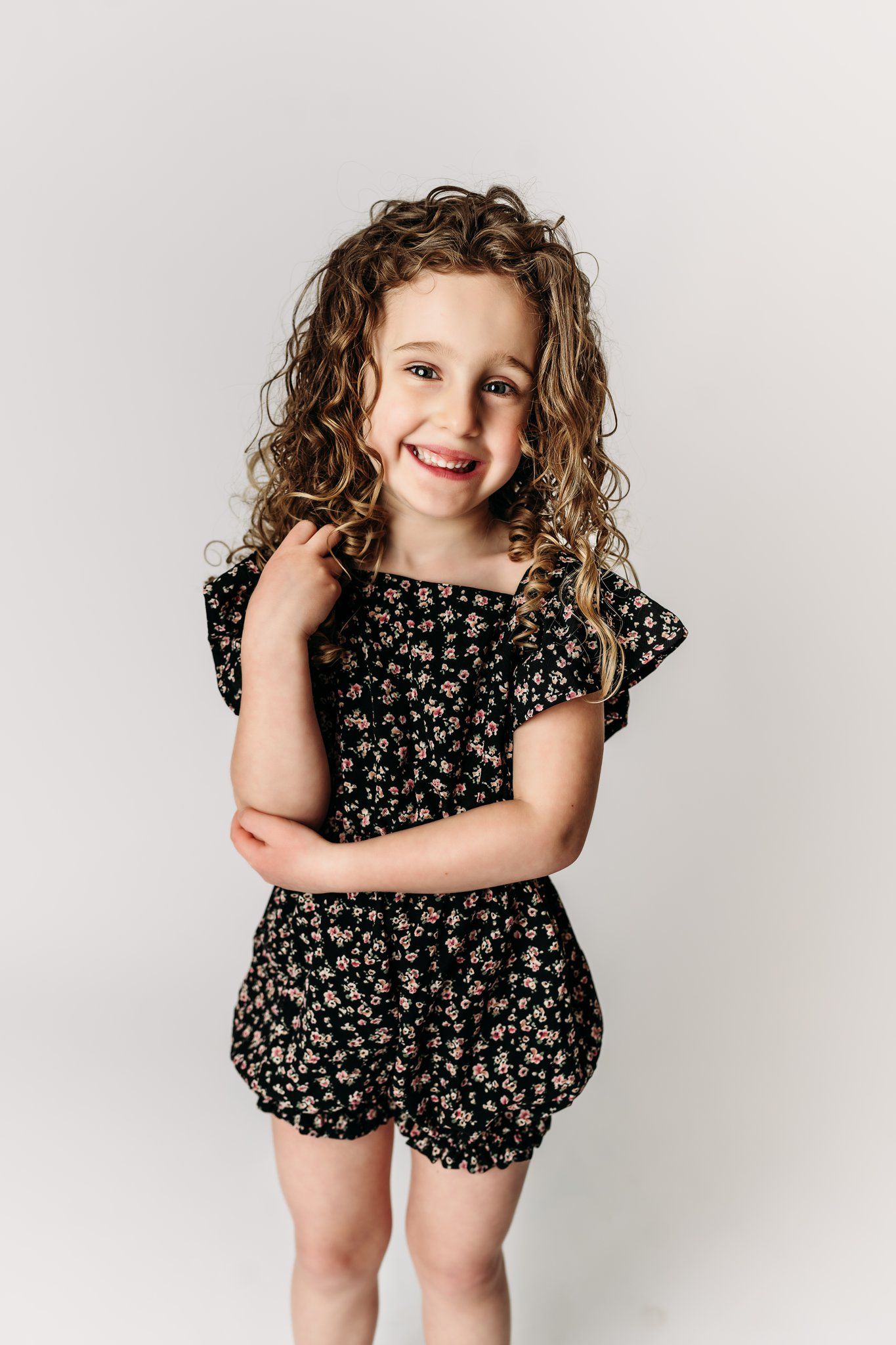 A child wearing a floral romper poses with crossed arms and a bright smile.