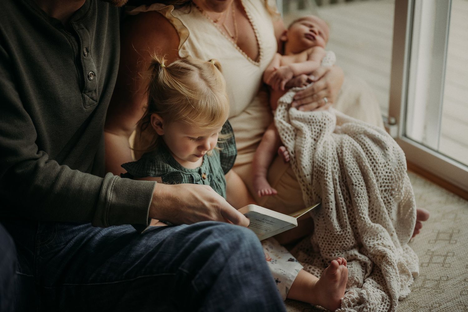 Family reading moment with toddler and infant by bright window.