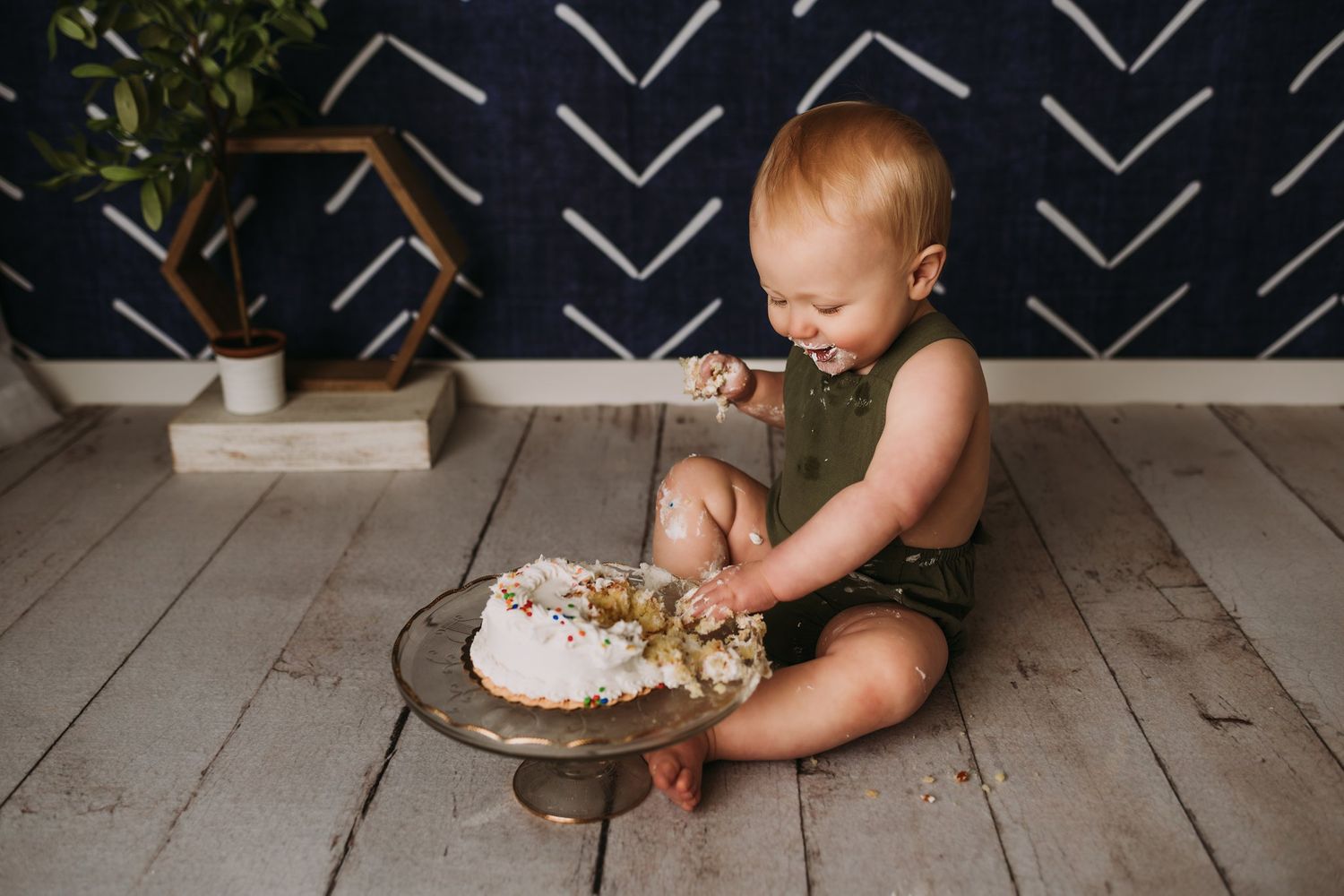 A final cake smash photo showing a demolished dessert on glass pedestal against navy patterned wall.