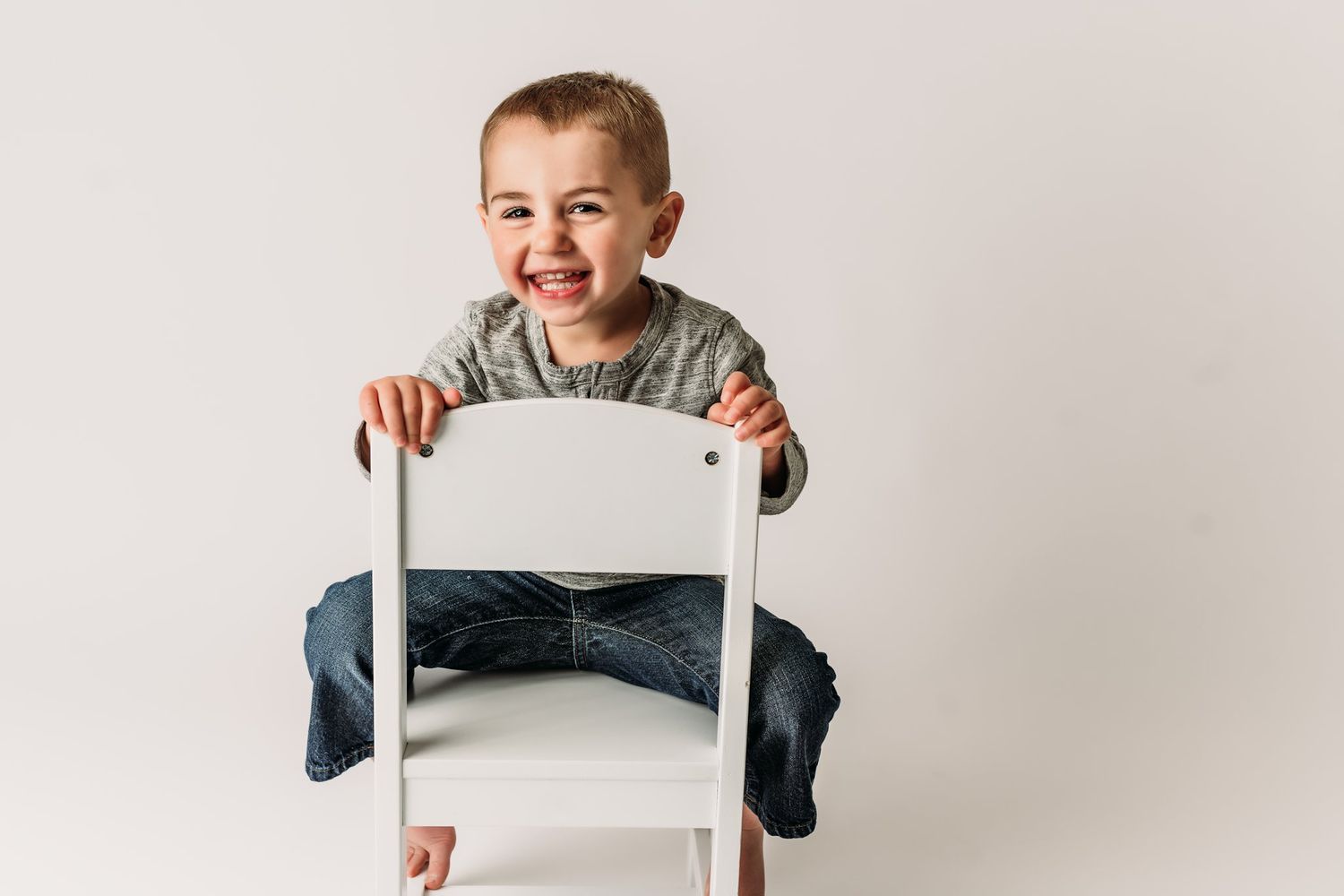 A joyful expression while sitting backwards on a white chair wearing a gray sweater and jeans.