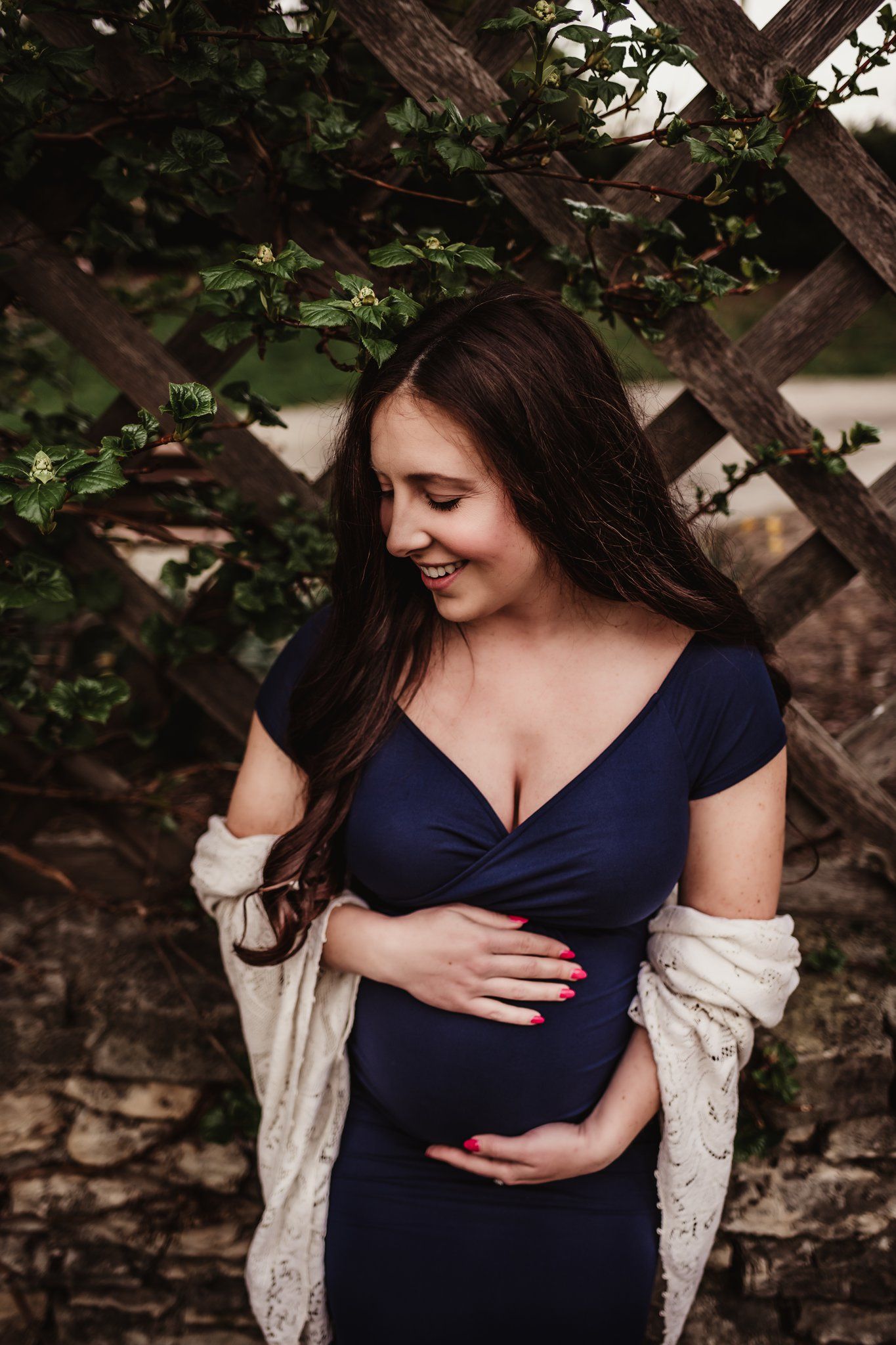 Expectant woman in navy dress and cream cardigan poses against rustic lattice wall with greenery.