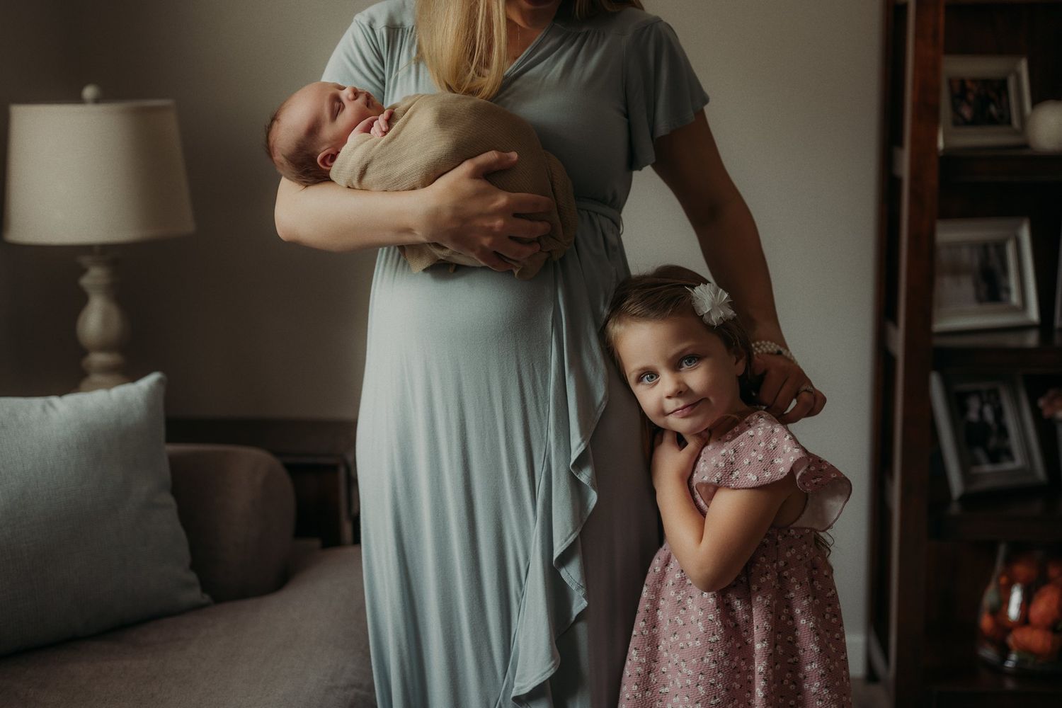 Family moment with parent holding newborn while child in pink dress stands nearby.