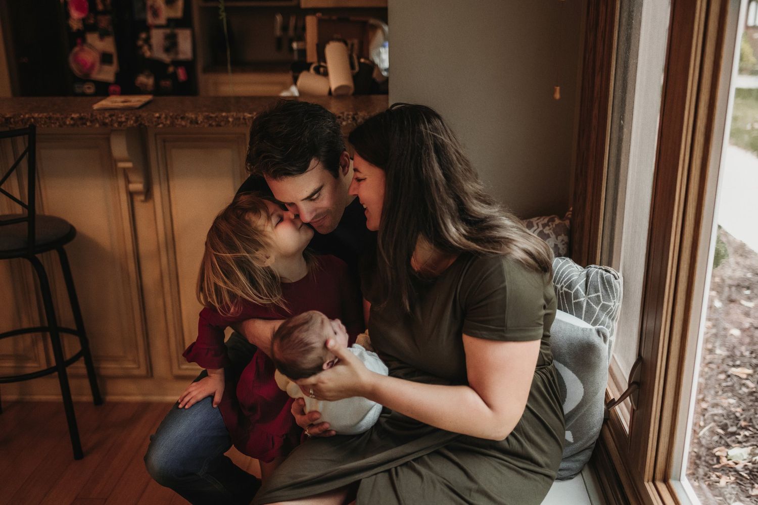 Tender family moment captured near a window in a warm, sunlit living room.