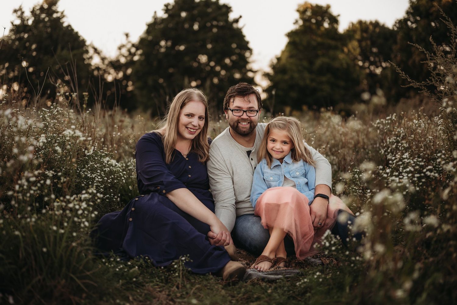 Family of three sitting together in a meadow during golden hour photography session.