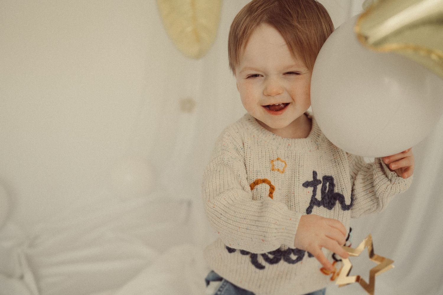Child smiling brightly in beige sweater against white backdrop with gold decorations.