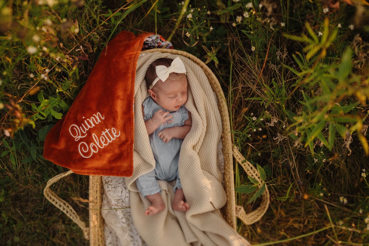 Newborn in light blue outfit resting in cream-colored Moses basket with red heart pillow in outdoor garden setting.
