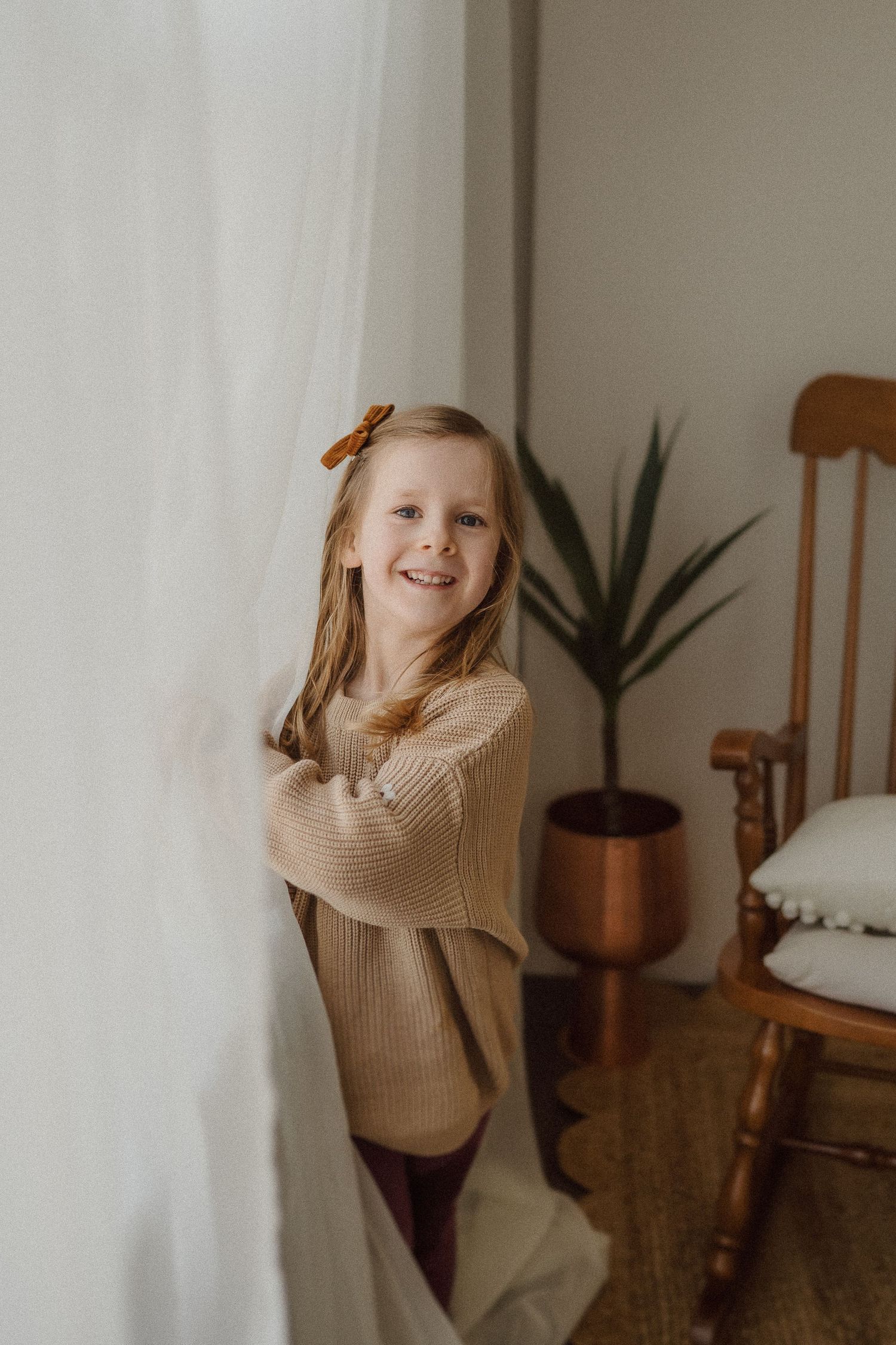 Natural light portrait by white wall with wooden chair and copper planters in background.
