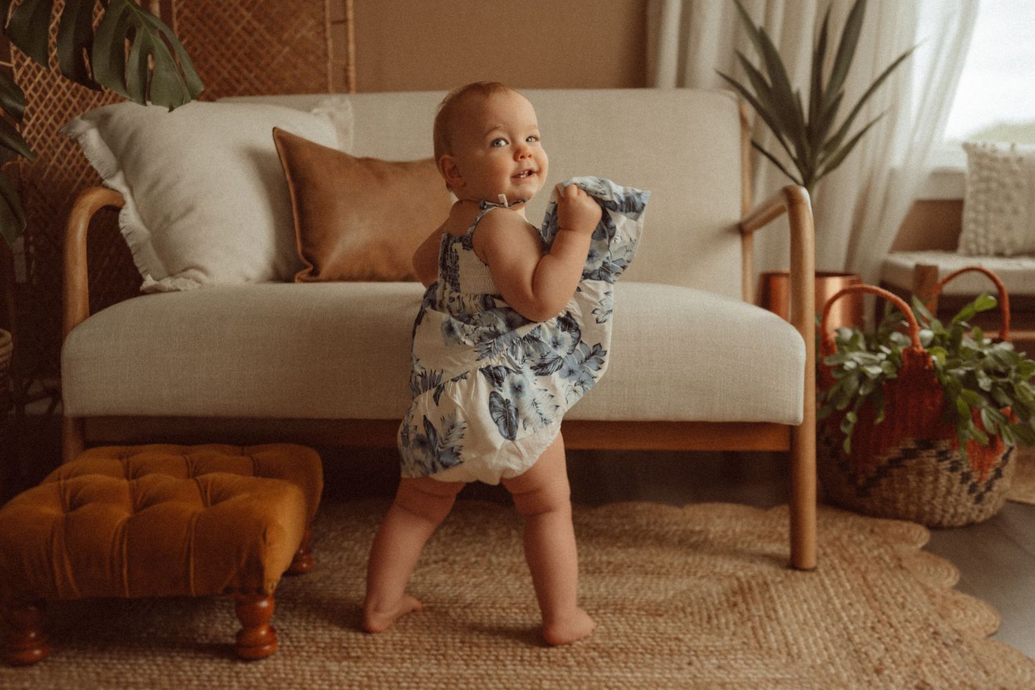 A playful toddler in a blue and white dress stands near a tan ottoman.